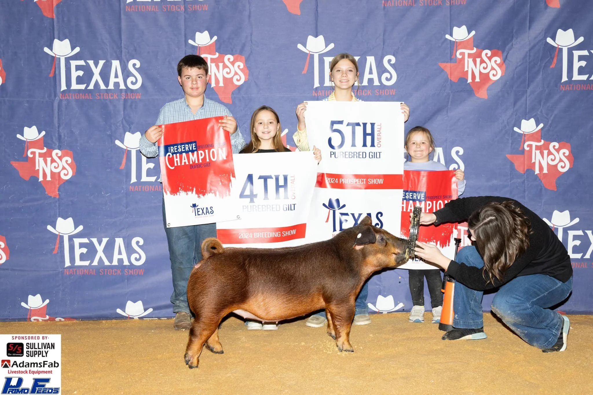 Children holding award signs and a woman with a pig at a livestock show with a Texas backdrop, showcasing awards for 4th and 5th place. The woman is feeding the pig.