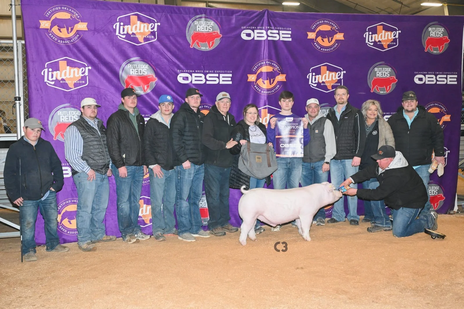 Group of people standing in front of a purple backdrop with logos, with a pig in the foreground at a livestock show. The people are posing, some holding a banner, and one kneeling, touching the pig's snout.