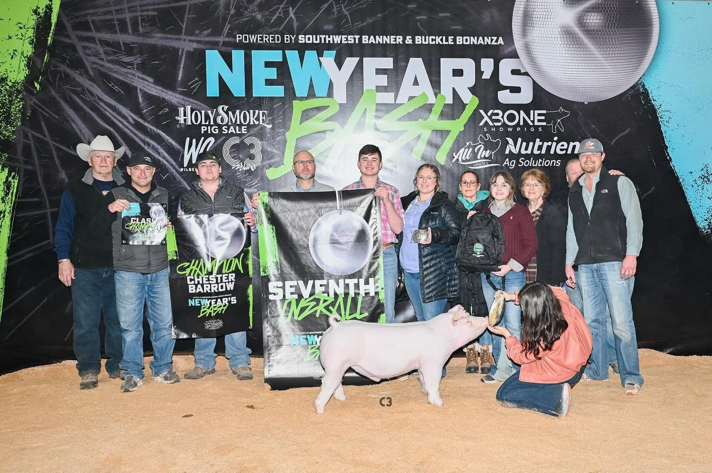 Group of people at a pig judging event holding banners, with a pig and a person feeding it in the foreground, large event backdrop with sponsors and text reading 'New Year’s Bash'.