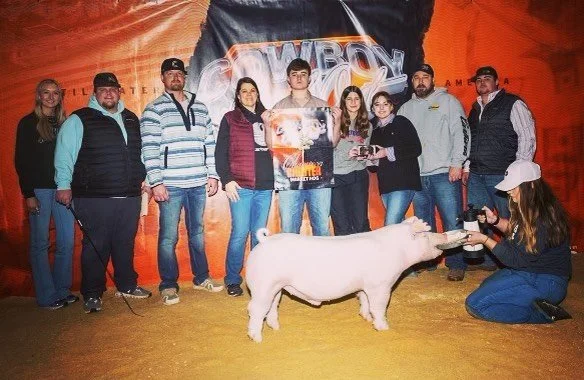 Group of nine people standing behind a pig at a livestock show or exhibition, with a woman taking photos of the pig.