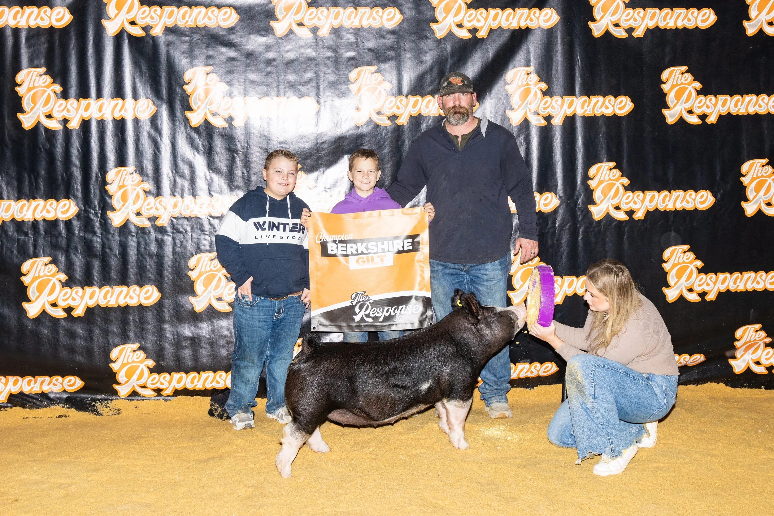 Group of four people, two children and two adults, at an event with a black backdrop that says 'The Response' in orange and white. The two children are holding a banner that says 'Berkshire Gilt.' One of the women is feeding a pig a treat, and there is a pig in the foreground, all on a yellow floor.