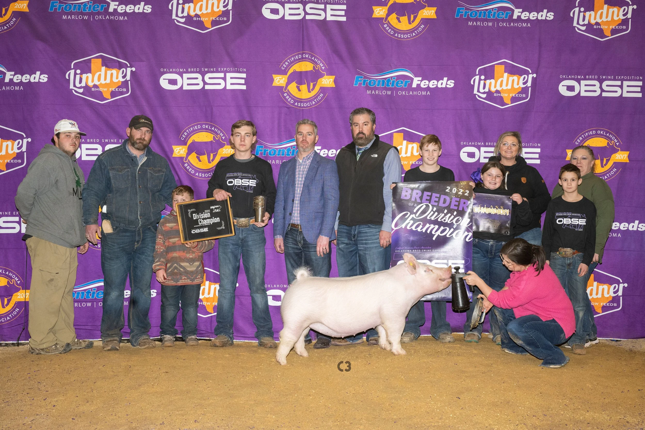 Group of people standing behind a pig at an Oklahoma bred swine exposition, with banners and signs indicating division champion and breeder of the year awards.