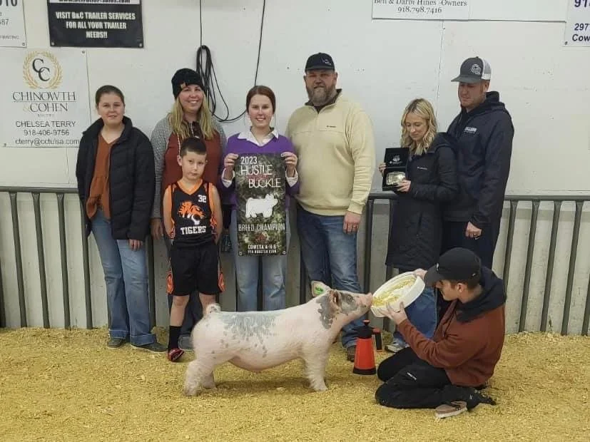Group of people at a livestock show with a pig. A woman holds a sign reading '2023 Hustle Buckle Breed Champion,' while a young man feeds the pig a bowl of food. The setting appears to be an indoor livestock arena with a metal railing and straw-covered ground.