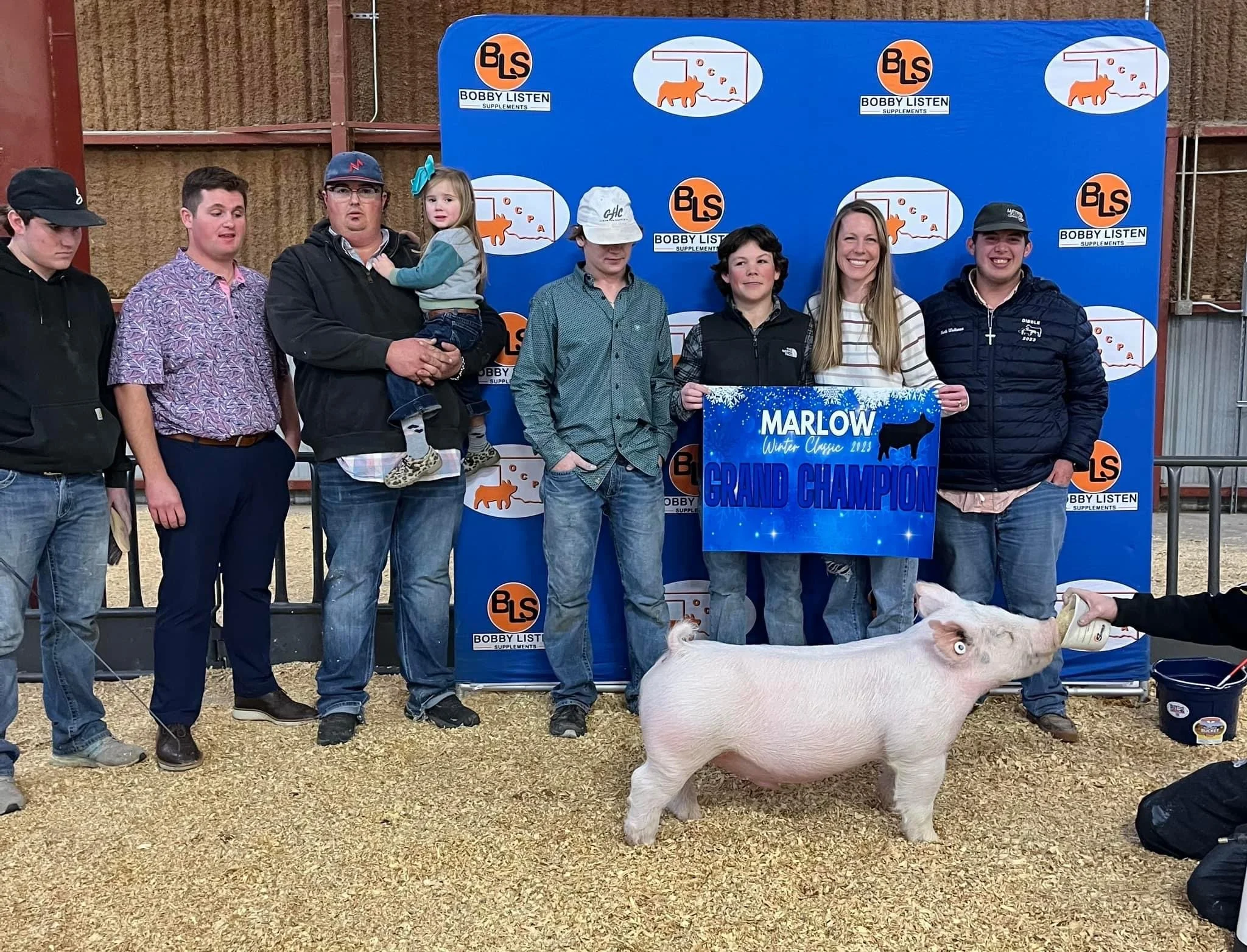 Group of people standing in a livestock exhibition with a pig in the foreground, holding a 'Grand Champion' sign for Marlow Winter Classic 2023, blue backdrop with logos and farm-themed images.