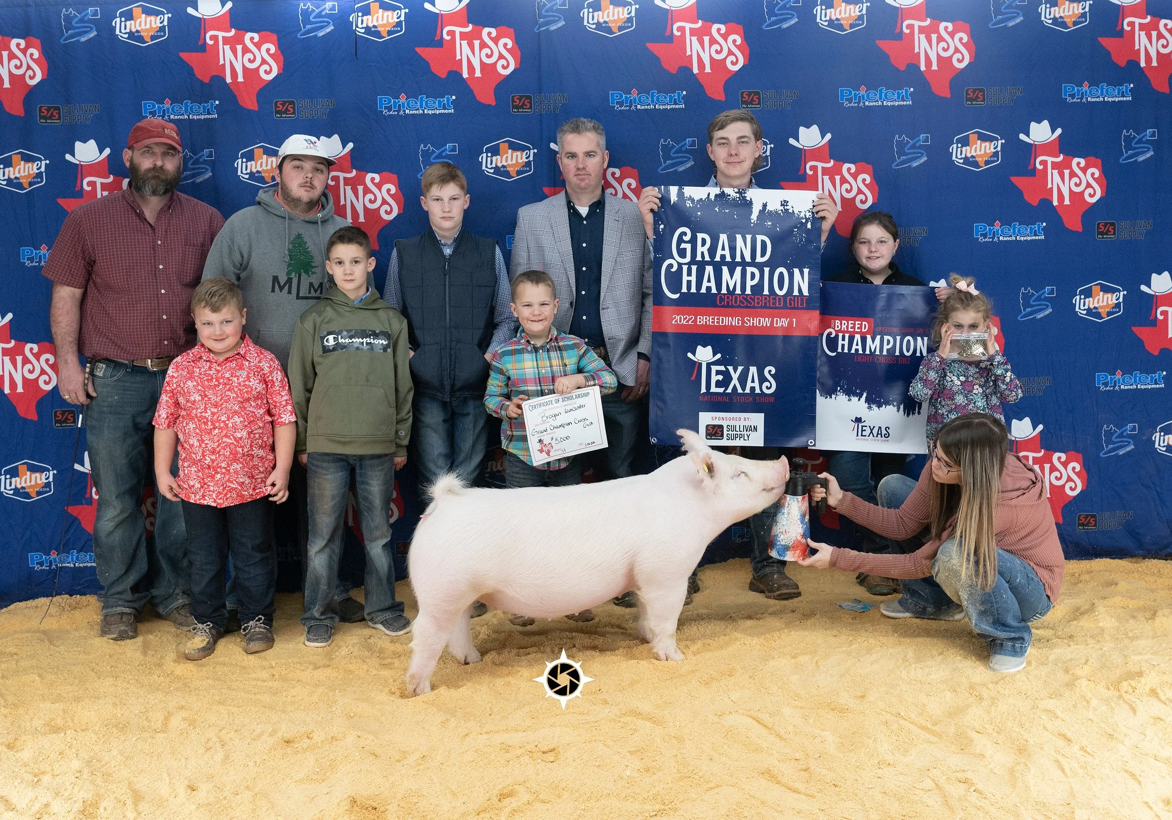 A group of people at a livestock show, with a pig in the foreground, holding banners that read 'Grand Champion' and '2022 Breeding Show Day 1'. The background features a blue banner with print relating to the event.