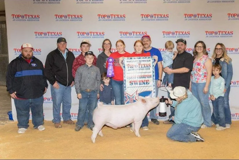 Group of people, including children, posing with a pig and a large winner's banner at the Junior Livestock Expo, with a backdrop displaying 'Top of Texas' and 'Junior Livestock Association' logos.