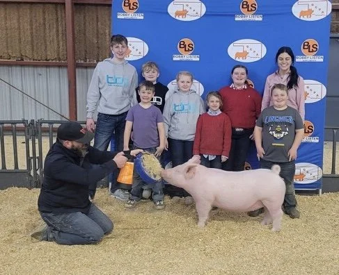 A group of seven children and two adults at a pig competition, with one man holding a paddling pool of feed and the pig standing on a dirt floor, in front of a blue and white backdrop.