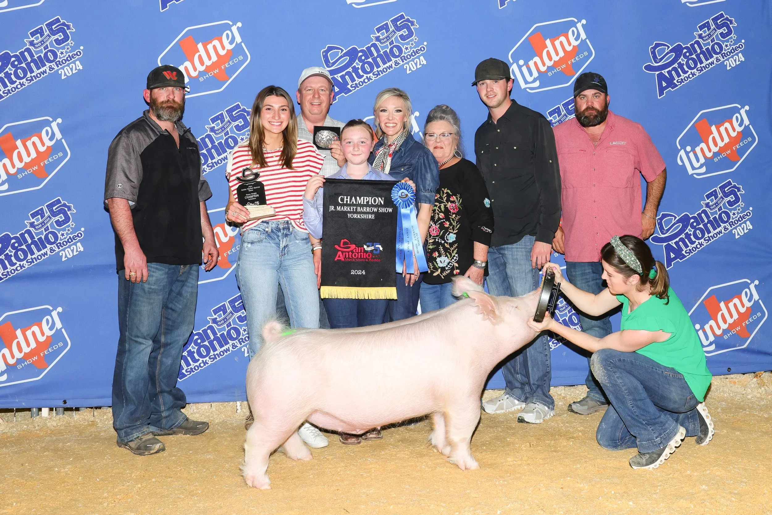 A group of people standing behind a large pig at an event, with a woman kneeling in front of the pig holding a small trophy. The background features a blue banner with the logos of San Antonio Stock Show & Rodeo 2024 and Lindner Show Feeds. The group includes men, women, and children, some smiling and holding awards, with the pig and a young girl in a green shirt in the foreground.