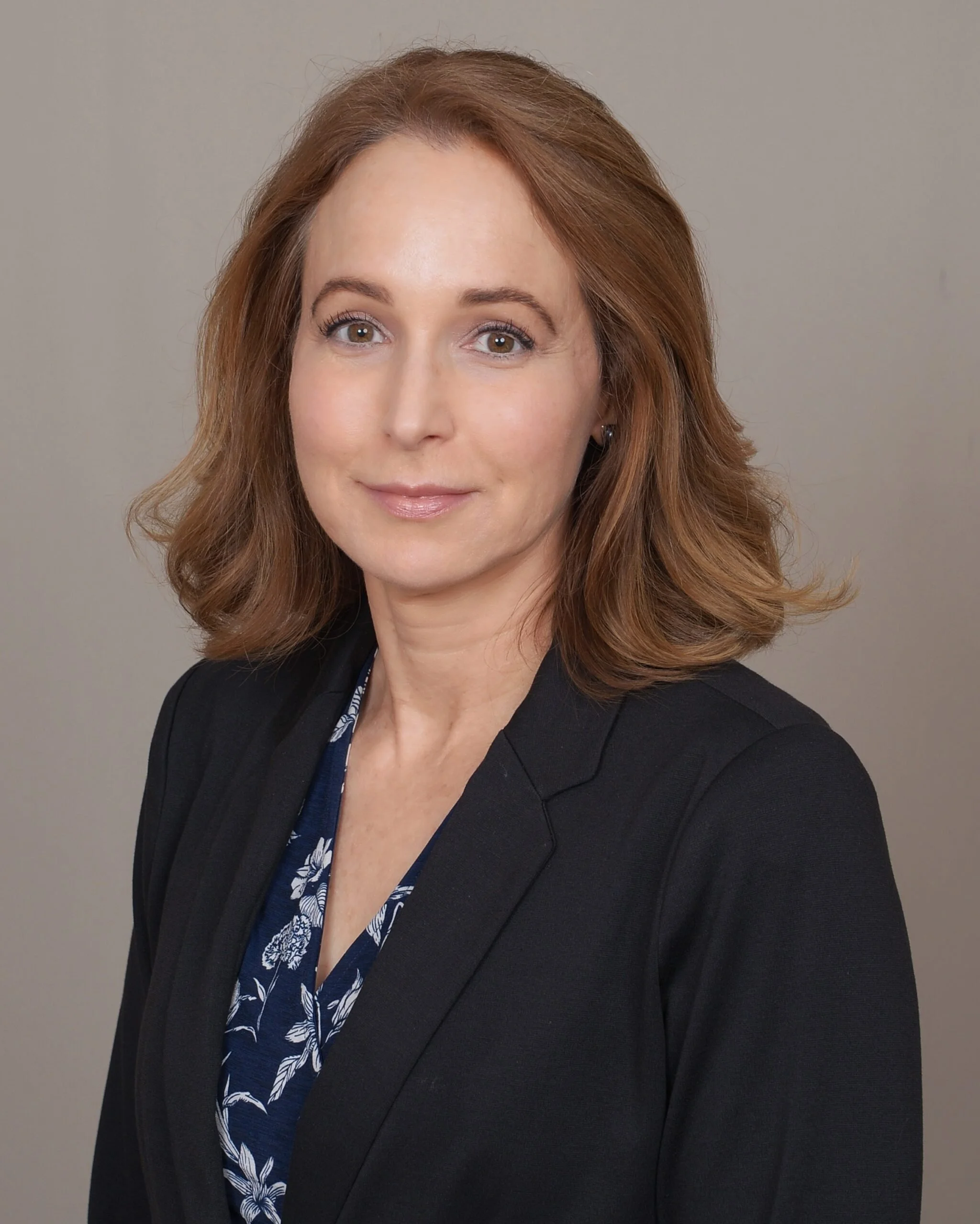 Headshot of a woman with shoulder-length red hair, wearing a black blazer over a blue patterned blouse, smiling softly against a neutral gray background.