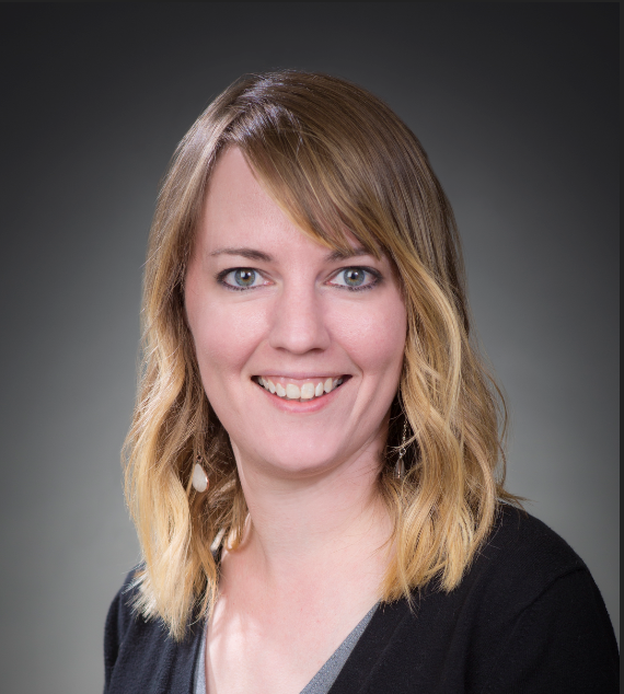 Headshot of a woman with shoulder-length blonde hair, blue eyes, smiling, wearing a black blazer and gray top, against a dark gradient background.
