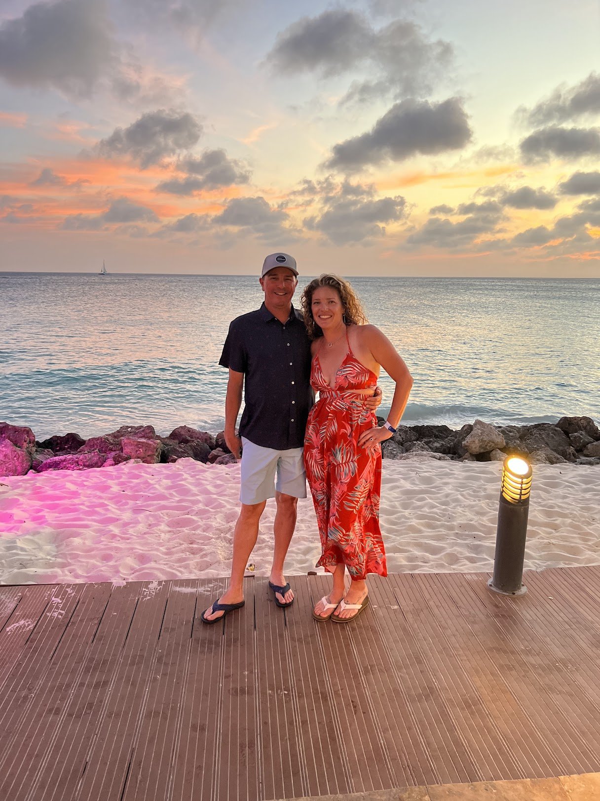 A man and woman standing on a wooden pier at sunset by the beach, smiling, with the ocean and cloudy sky in the background. Bookkeeping Services for small businesses virtually or in-person.