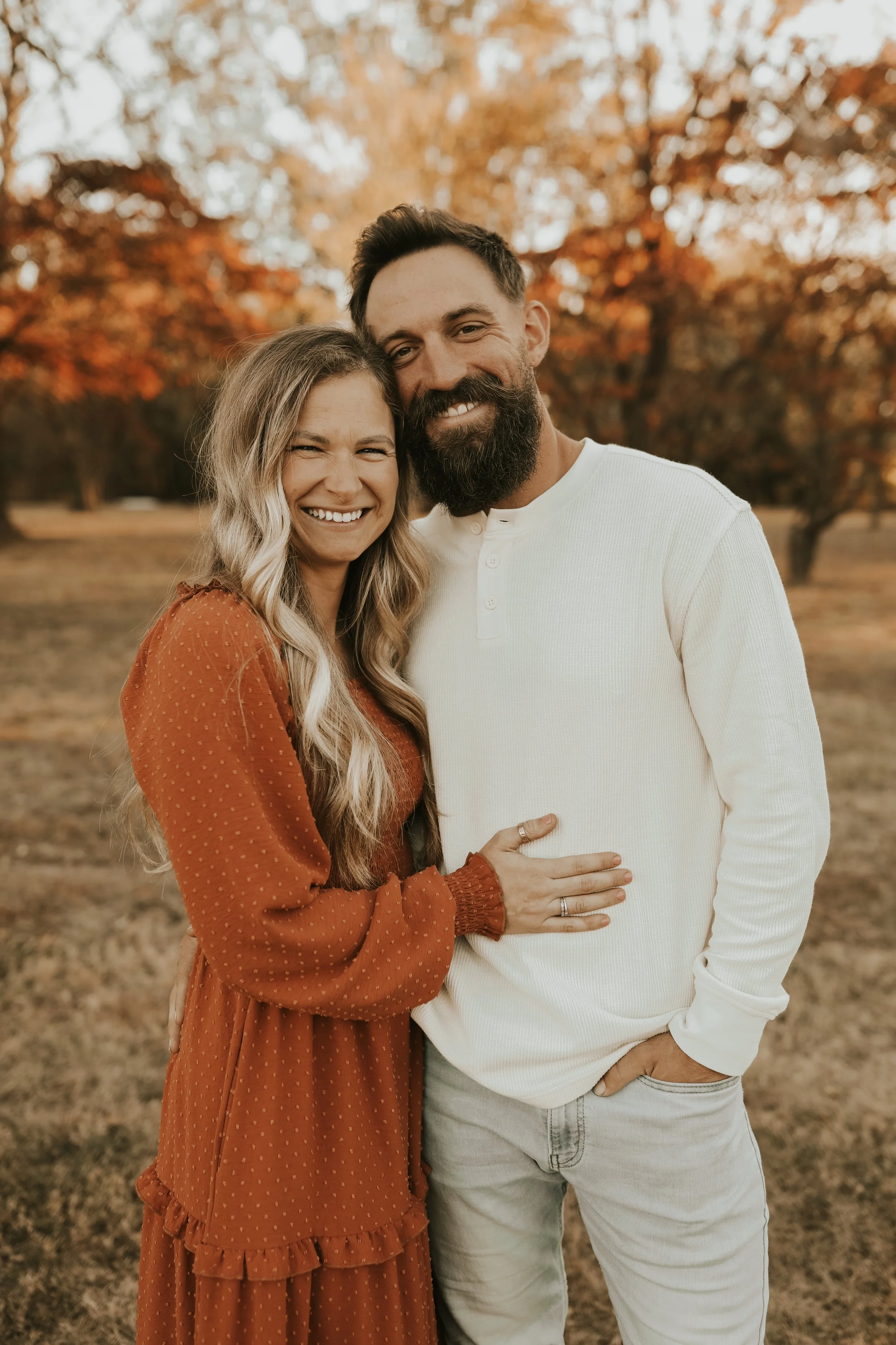 A happy couple standing outdoors in front of fall trees, smiling and embracing each other.