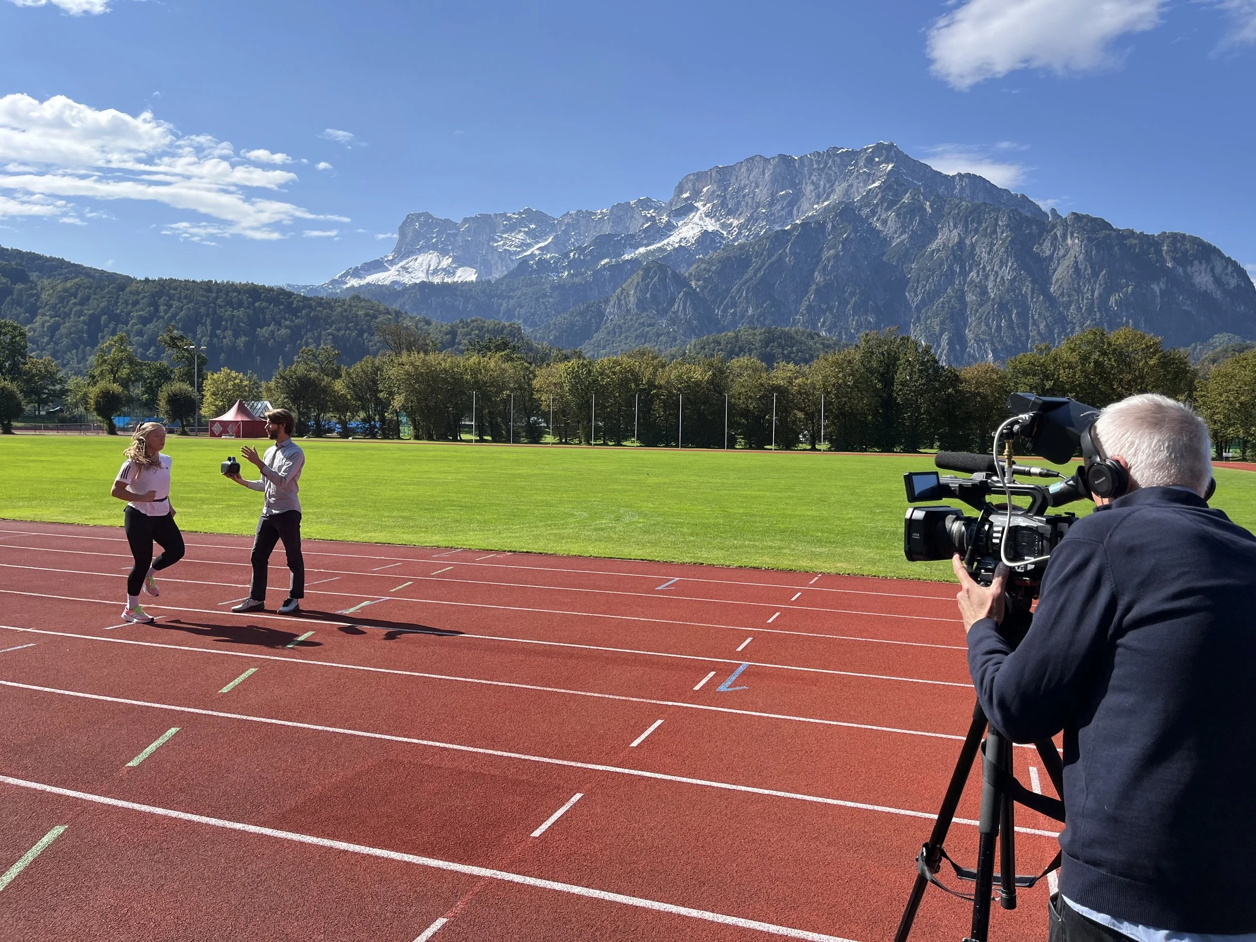 A man and woman running on a track while being filmed or photographed by a cameraman with a large camera, with green grass, trees, mountains, and a blue sky in the background.