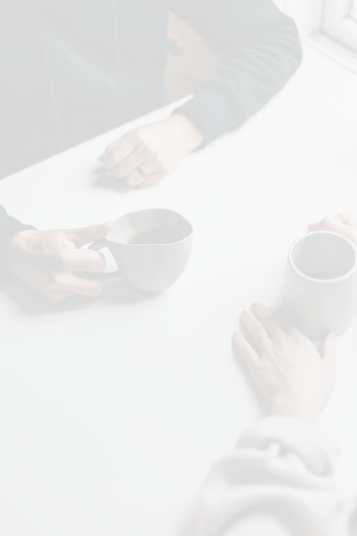 Blurred image of several hands reaching towards a table, holding cups of coffee, against a background of a window with bright natural light.