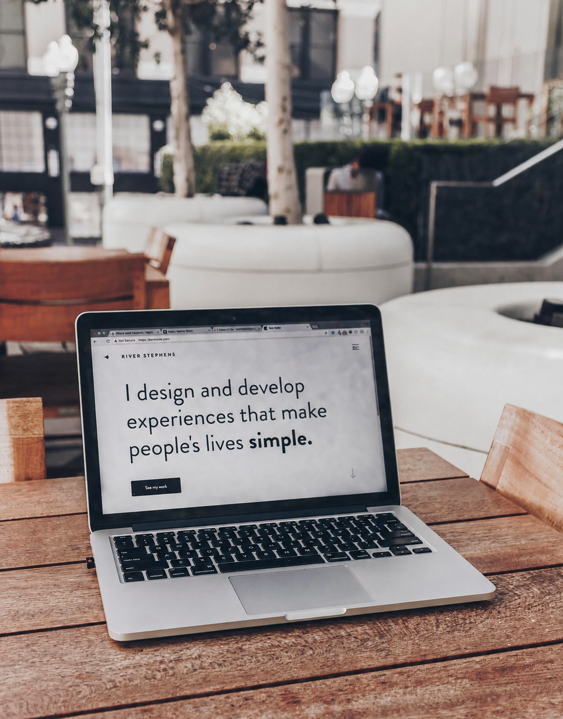 A laptop on a wooden table displaying a webpage with a quote about designing and developing experiences that make people's lives simple, in an outdoor cafe setting with trees and seating in the background.