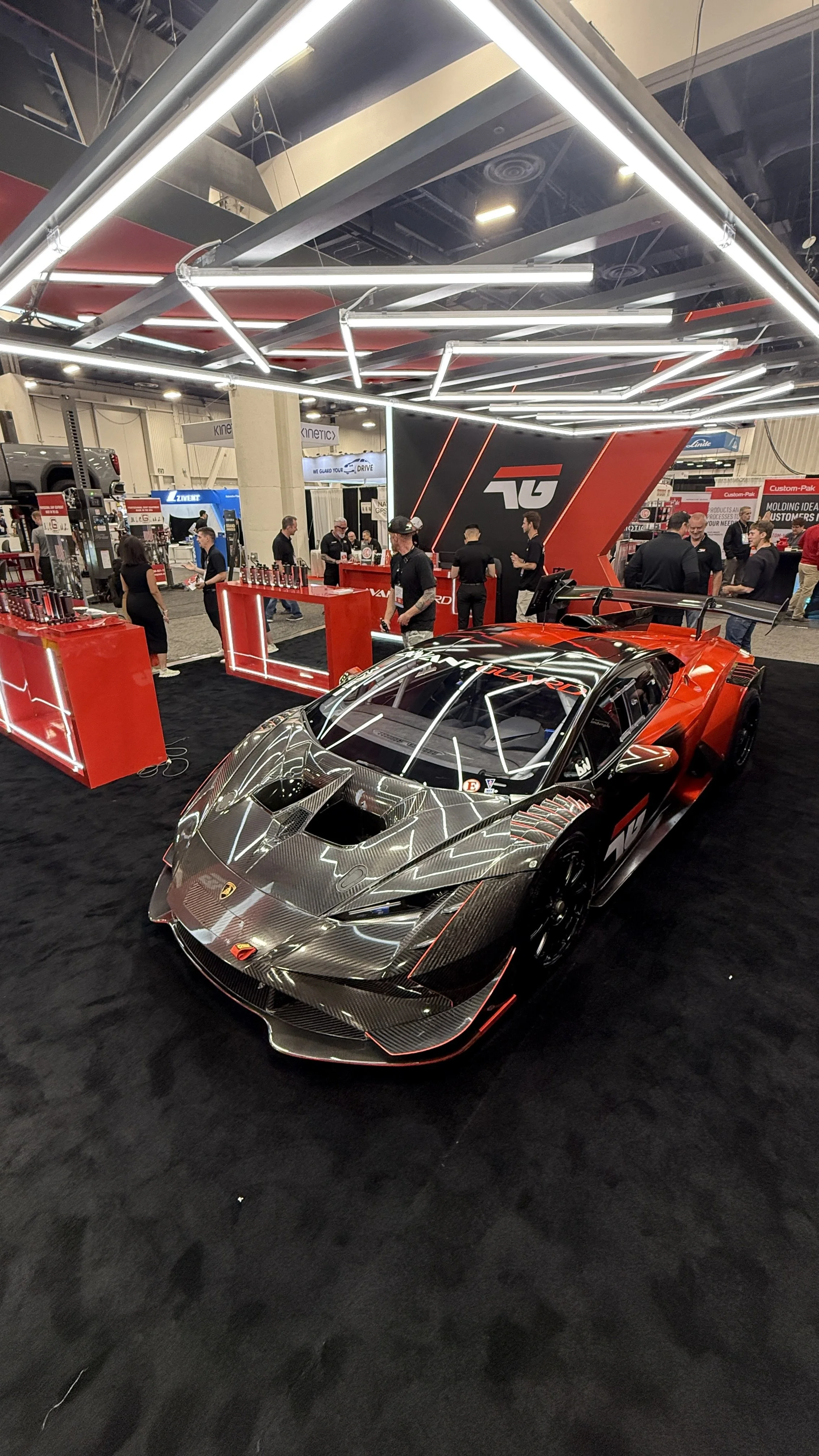 A Lamborghini race car on display at an auto show, with people in the background and a large TAG logo on the wall behind it.