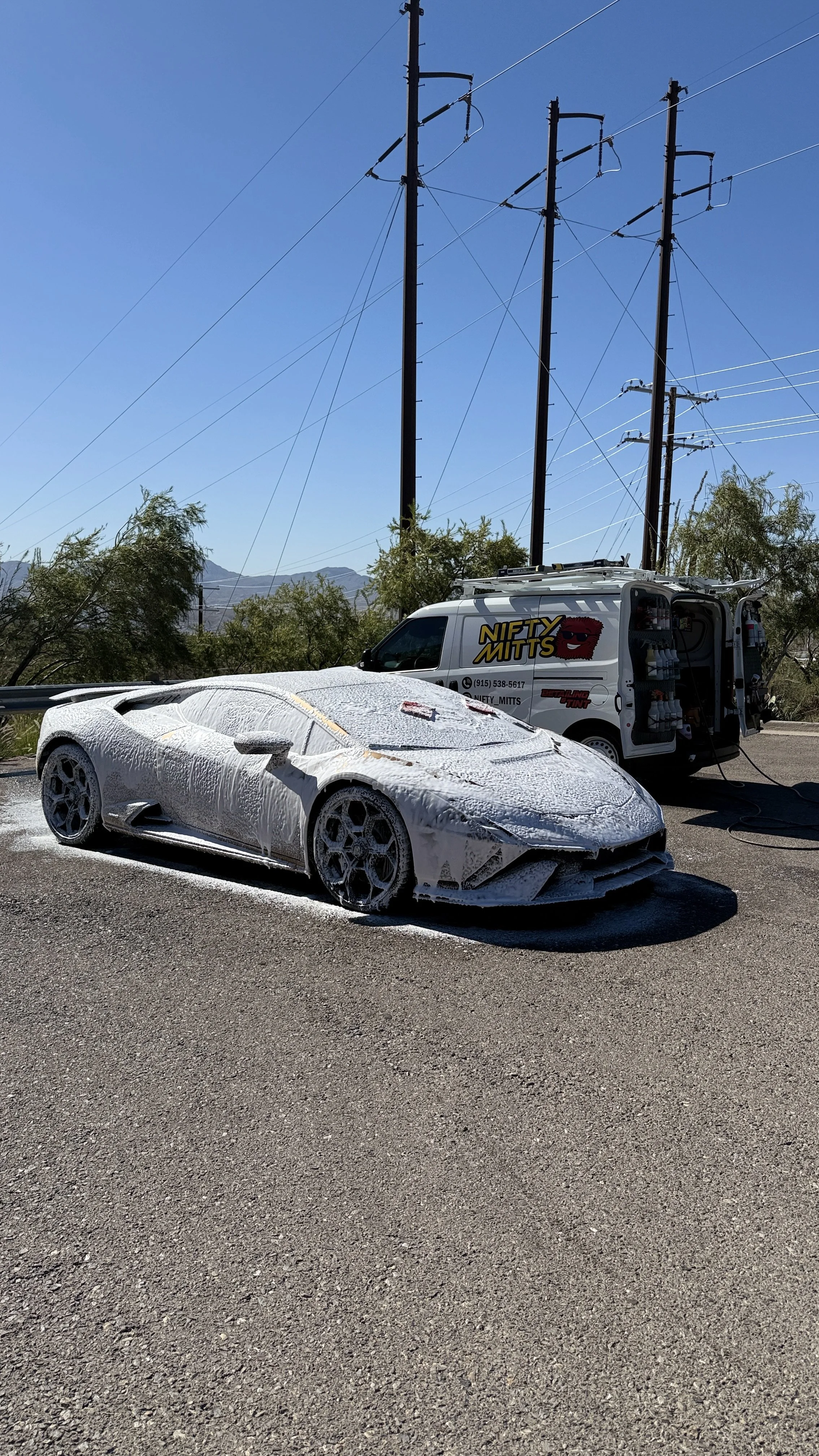 A white sports car covered in foam during a foam wash process, parked on an asphalt road background with utility poles and a service van labeled 'Nifty Mitts'.