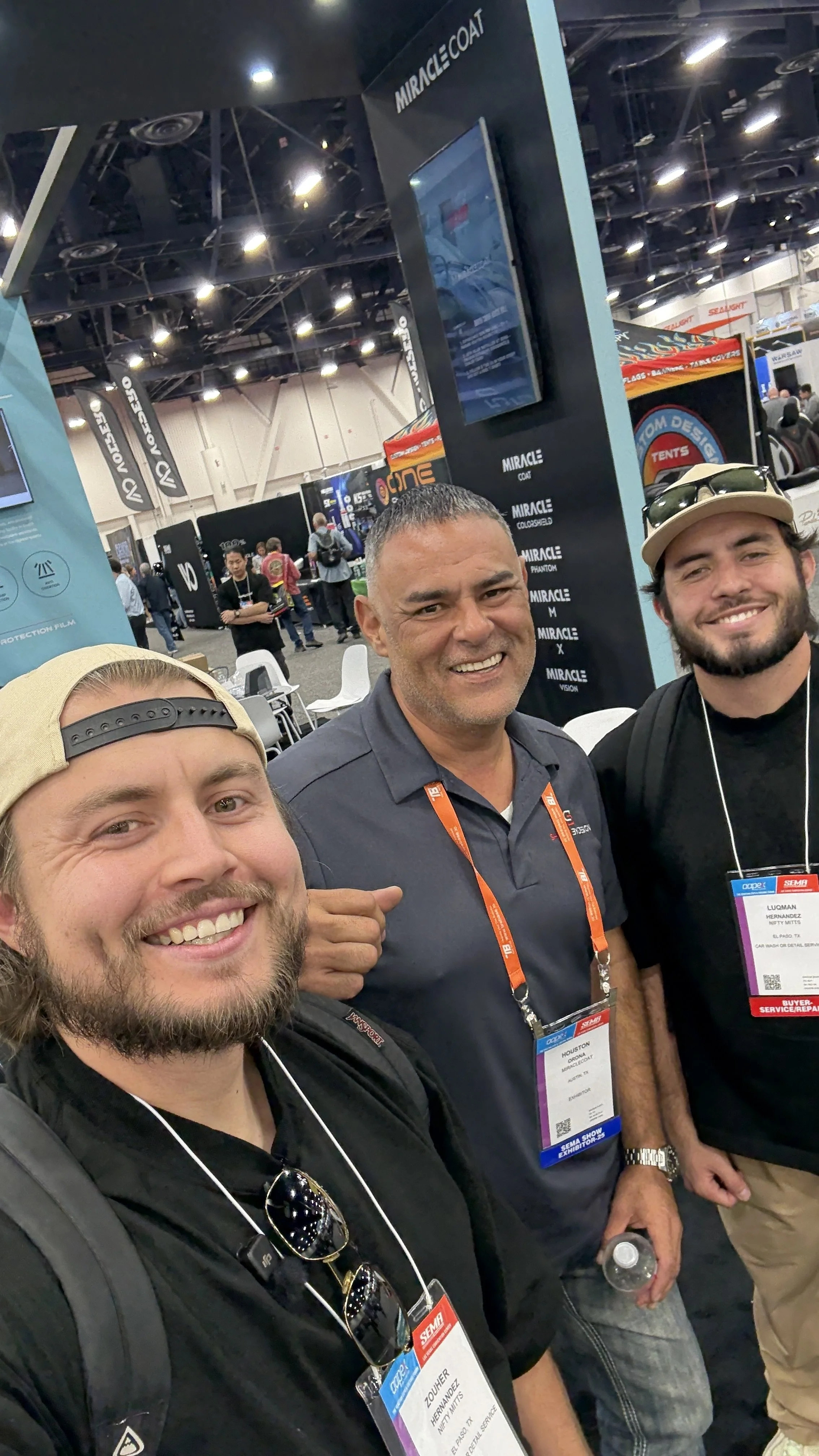 Three men smiling at a trade show or convention, with booths and other attendees in the background. They are wearing name tags on lanyards around their necks.
