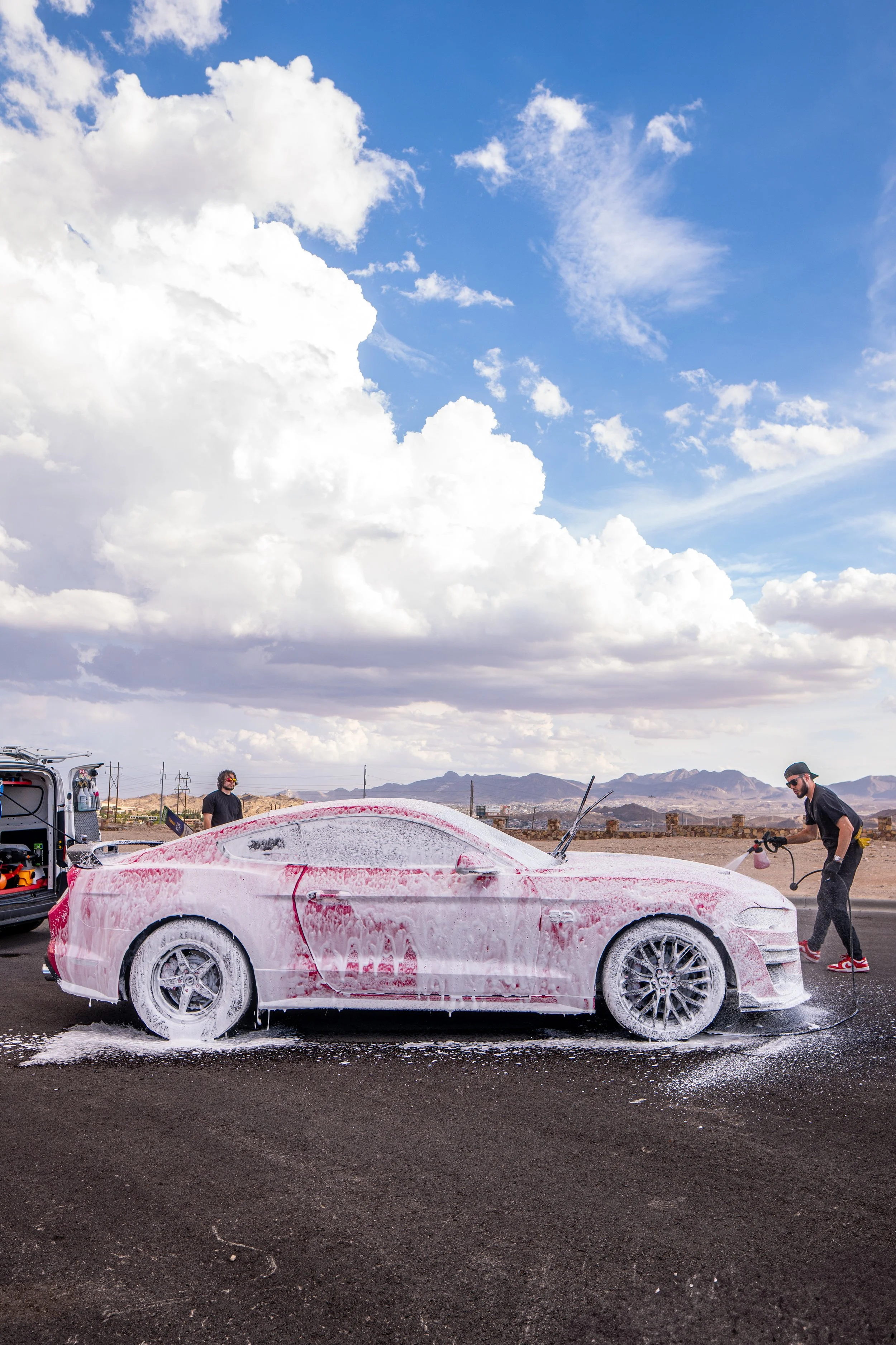 A person washing a pink sports car with soap foam on the side of a road in a desert landscape during daytime.