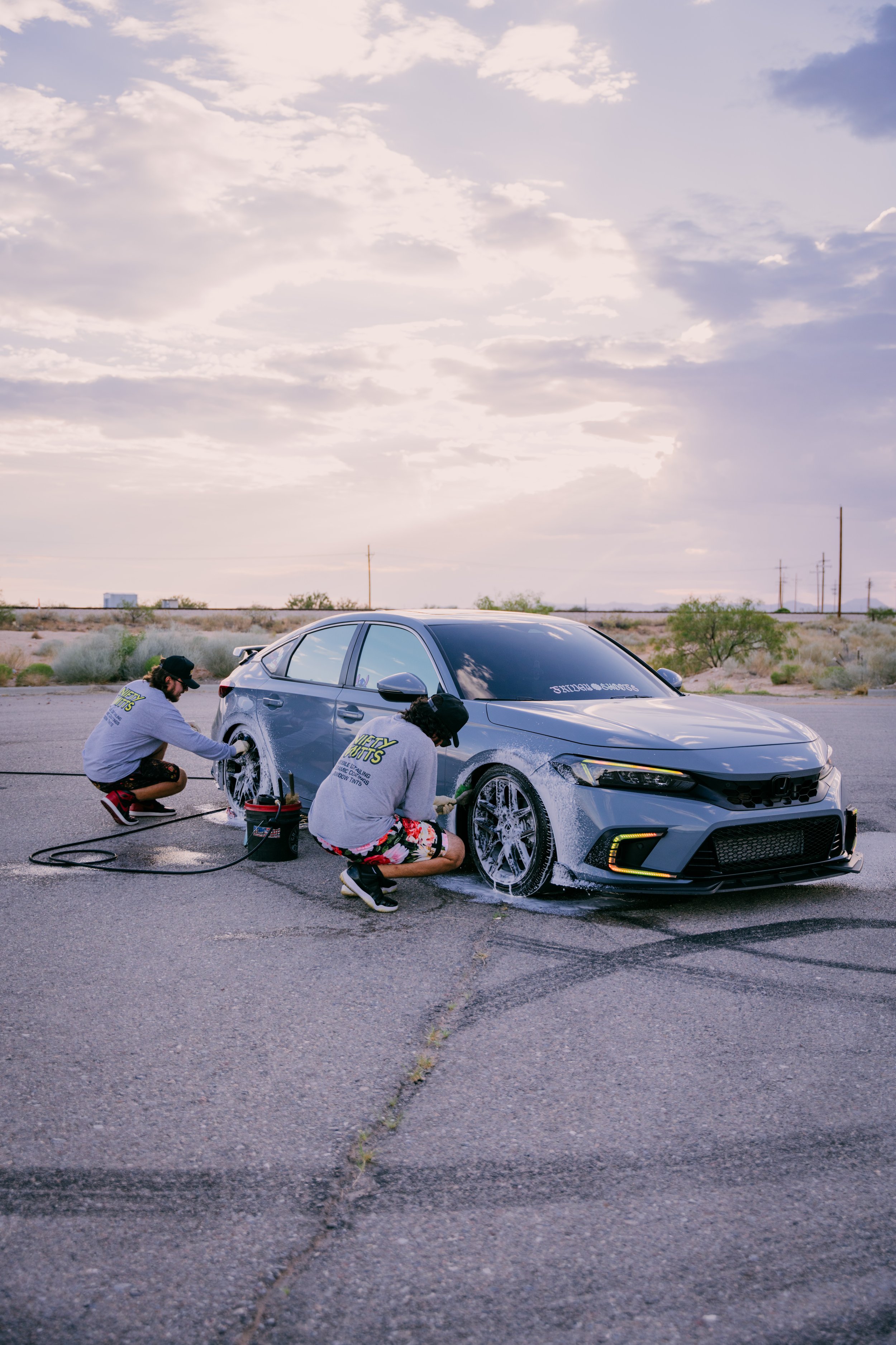 Two people washing a gray sports car in an open, desert-like area during sunset.