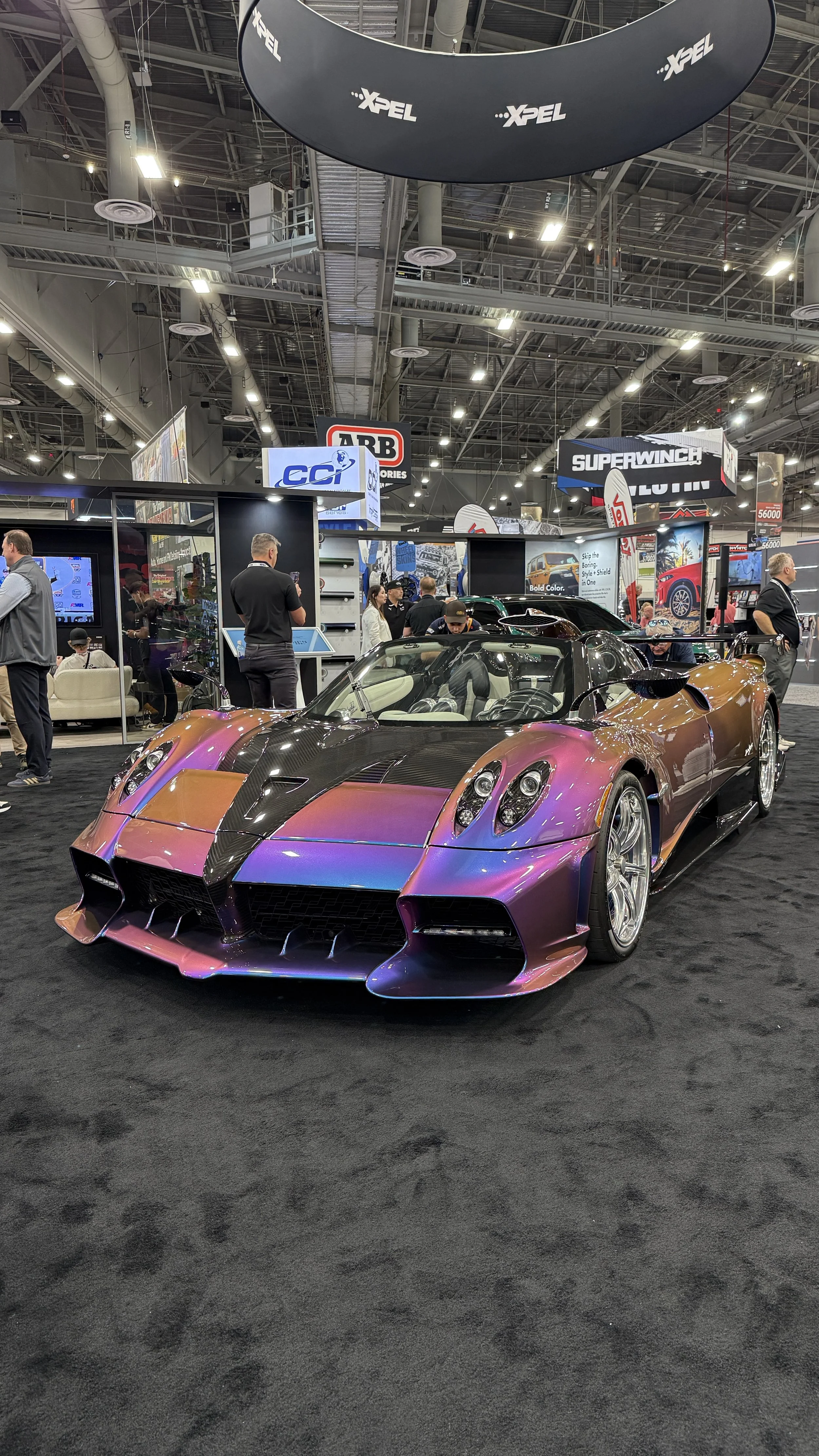 A multi-colored sports car with a low profile, large rear wing, and aerodynamic design at an auto show. The car has purple, gold, and black hues with racing-style wheels.