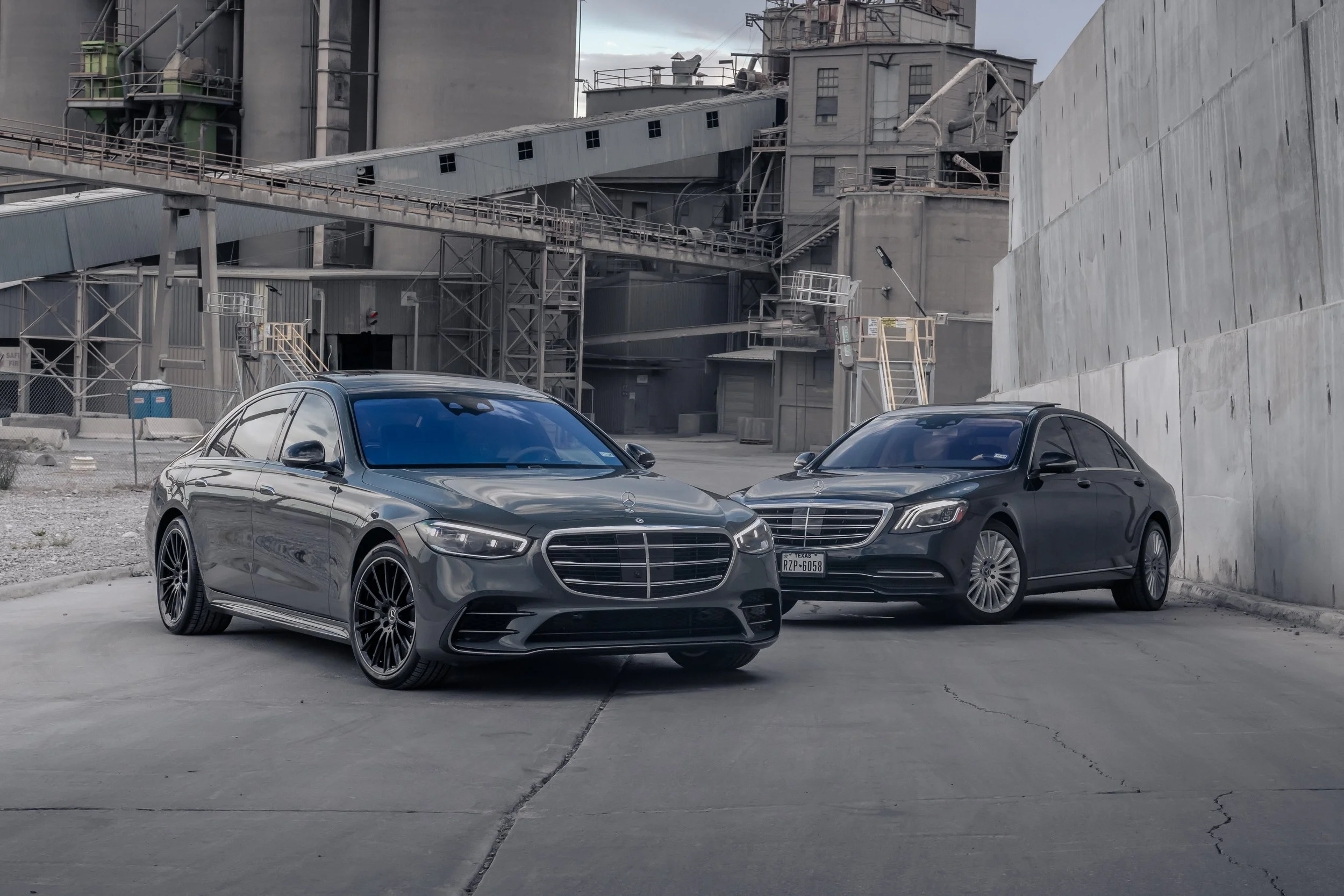 Two black Mercedes-Benz cars parked in an industrial, concrete environment with towering silos and metal staircases in the background.