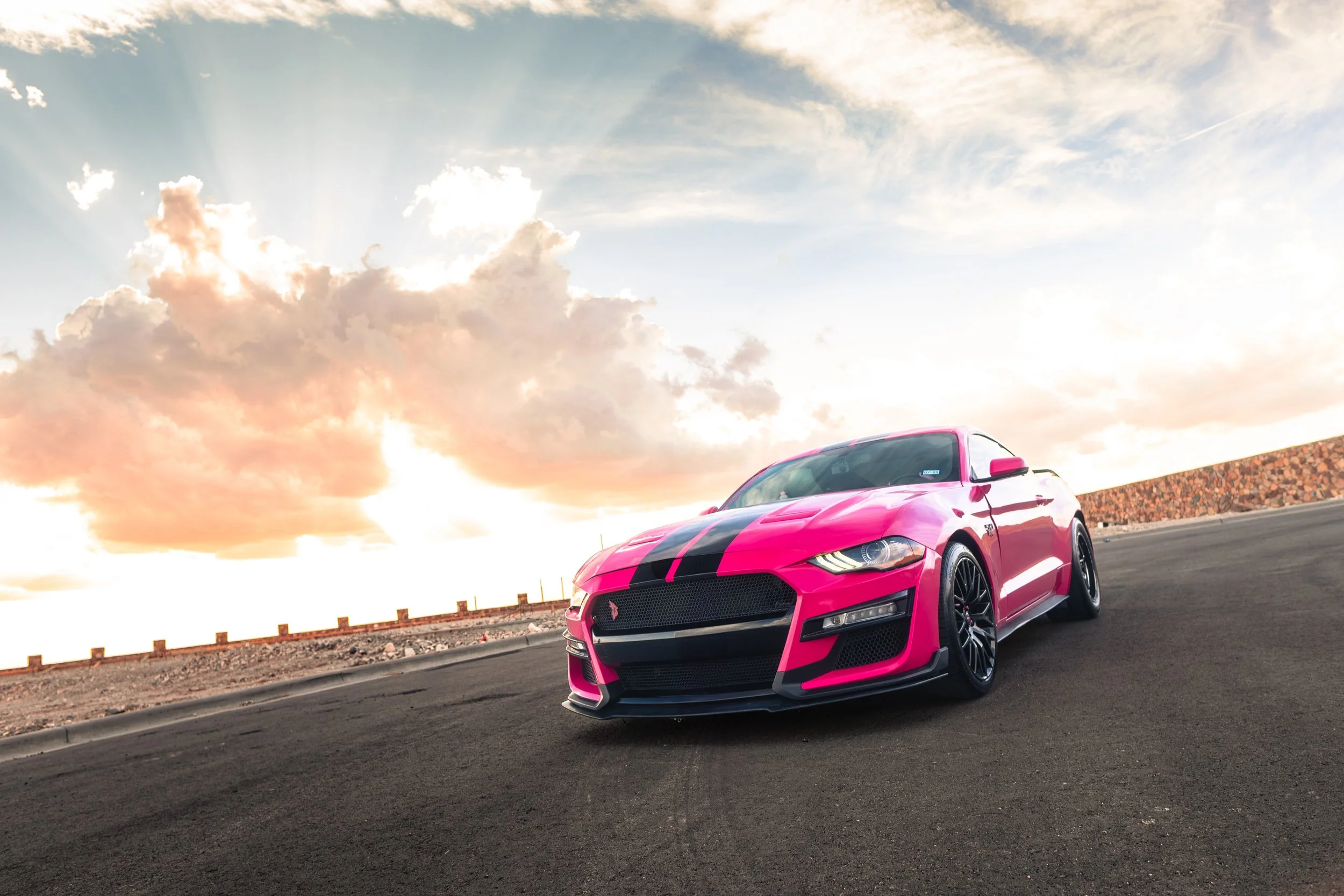 A pink sports car with black racing stripes parked on an empty asphalt road during sunset, with a cloudy sky overhead.