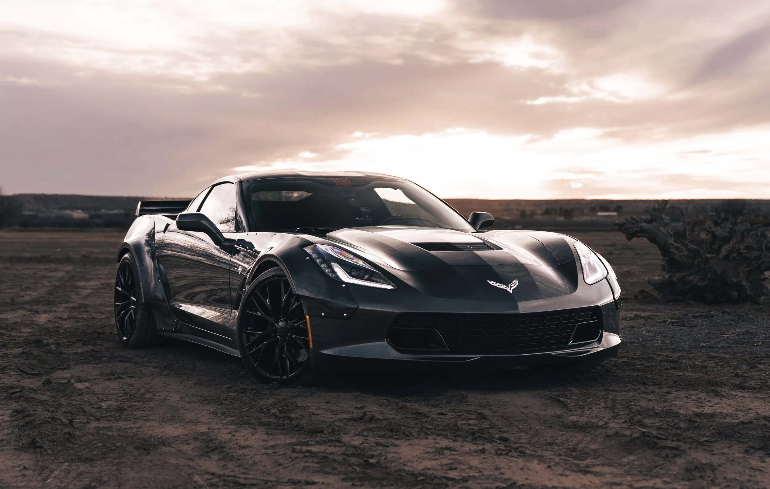 A black Chevrolet Corvette sports car parked on dirt ground at sunset with a cloudy sky and open landscape in the background.