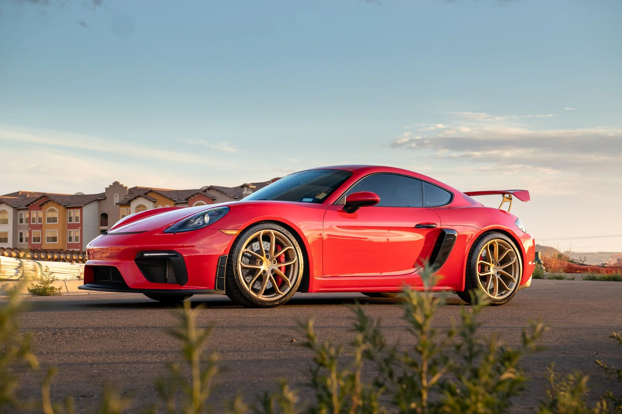 Red sports car with gold wheels parked on a street with residential buildings in the background during sunset.
