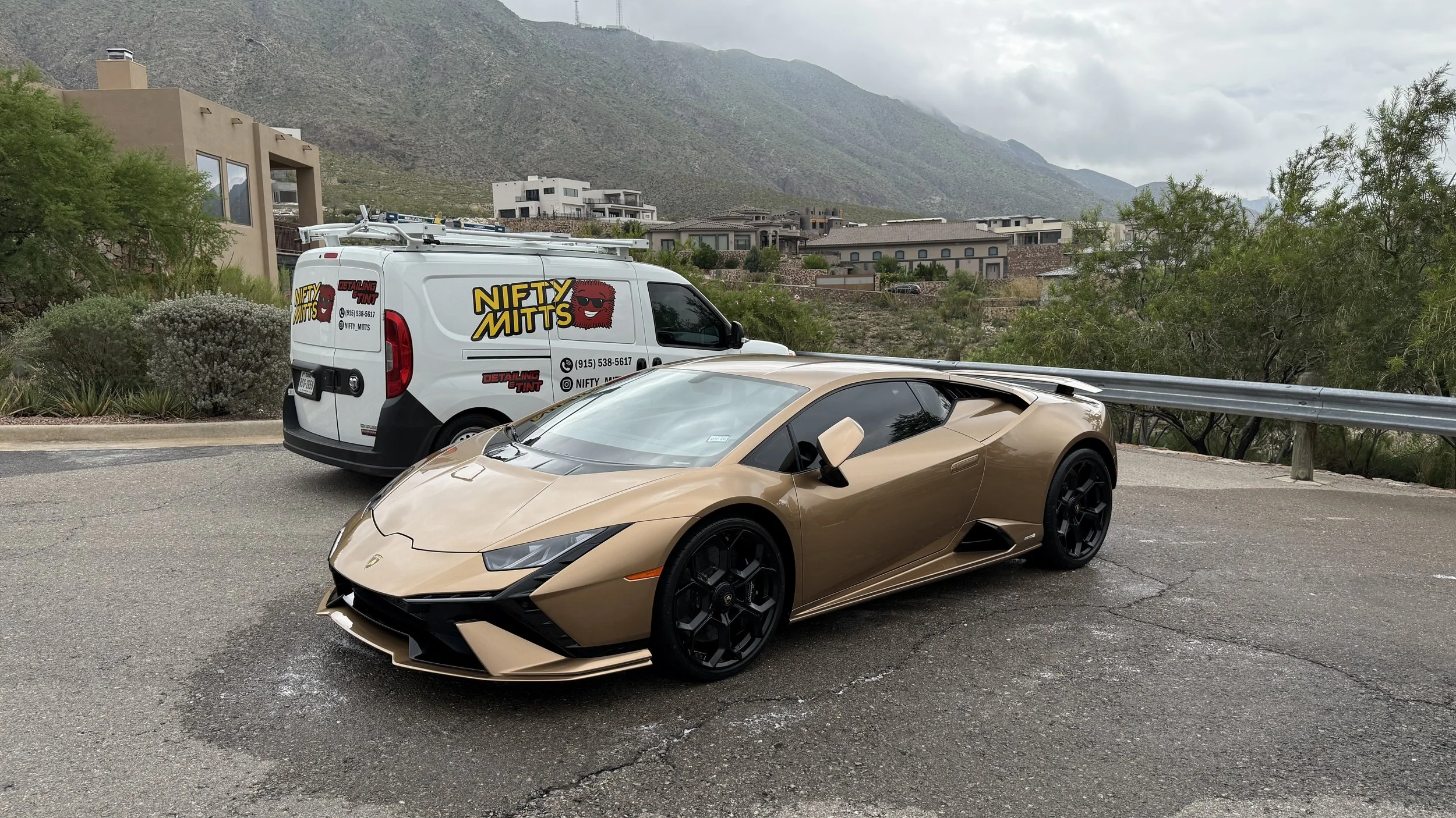 Gold Lamborghini sports car parked on the side of the road with a white vehicle marked 'Nifty Mitts' in the background, trees, and residential buildings under a cloudy sky.