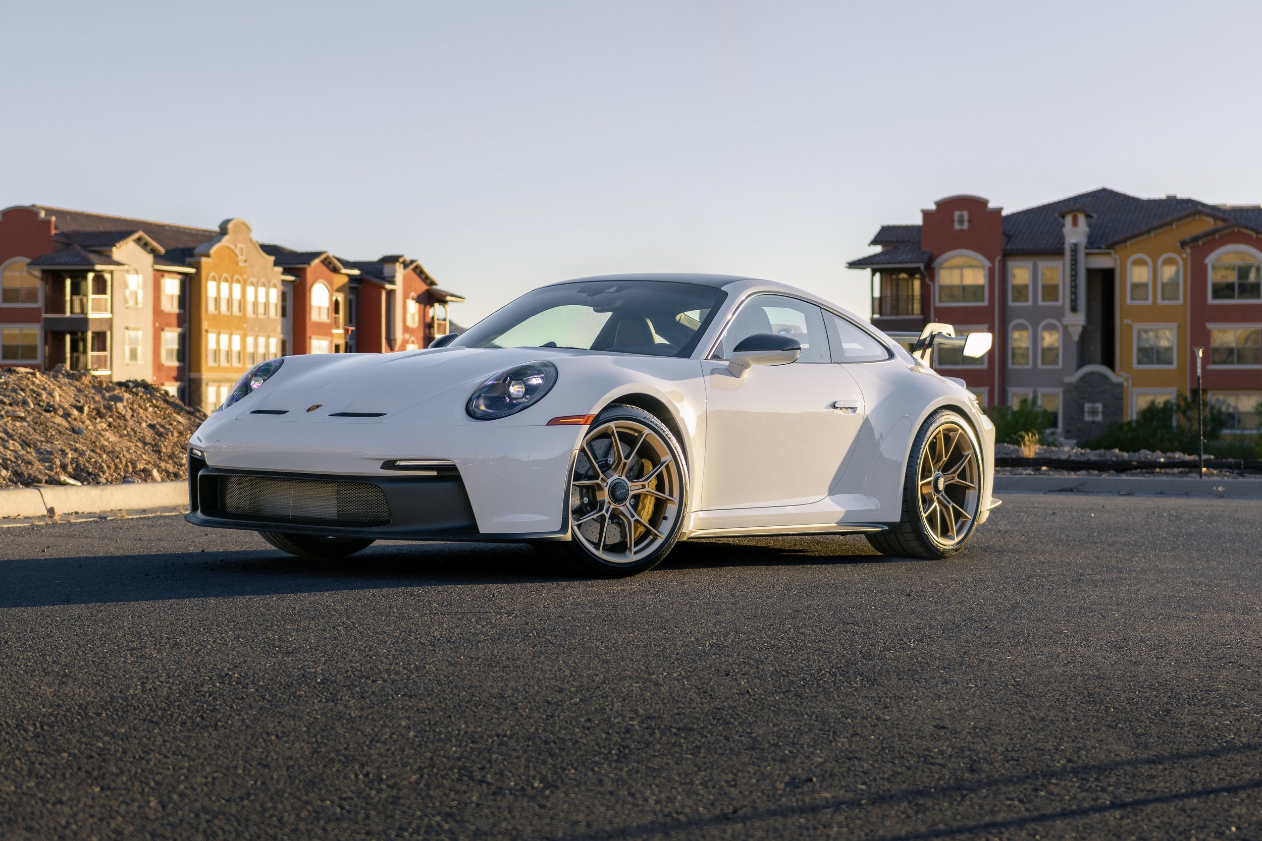 A white sports car with gold rims parked on a paved street in front of colorful residential buildings during sunset.