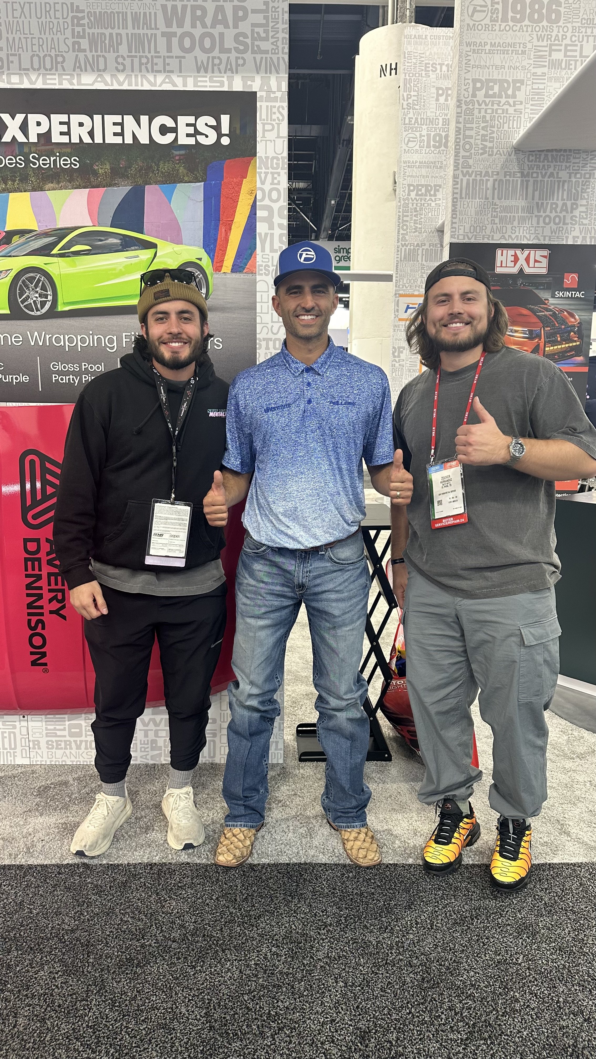 Three men standing indoors, smiling at the camera, giving thumbs-up gestures. The man on the left wears a black hoodie, beige beanie, and white sneakers. The man in the middle wears a blue patterned polo shirt, jeans, and snake skin boots. The man on the right wears a gray t-shirt, gray cargo pants, and tiger-striped sneakers. Behind them are promotional banners featuring cars and the text 'EXPERIENCES!' and 'EVERY DENNISON'.