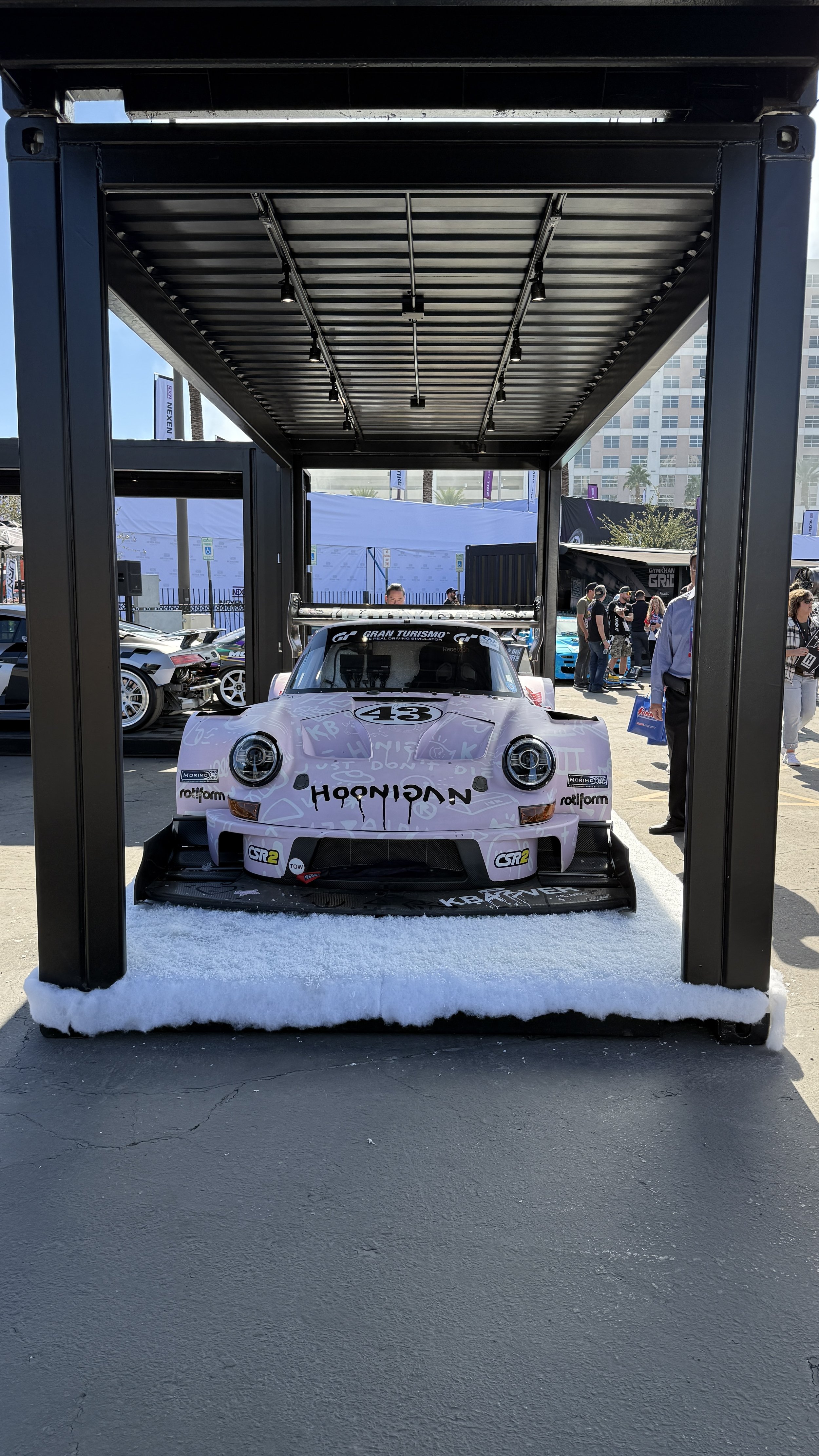 A race car displayed under a black structure with a snow-like base at an outdoor event, surrounded by people and other vehicles.