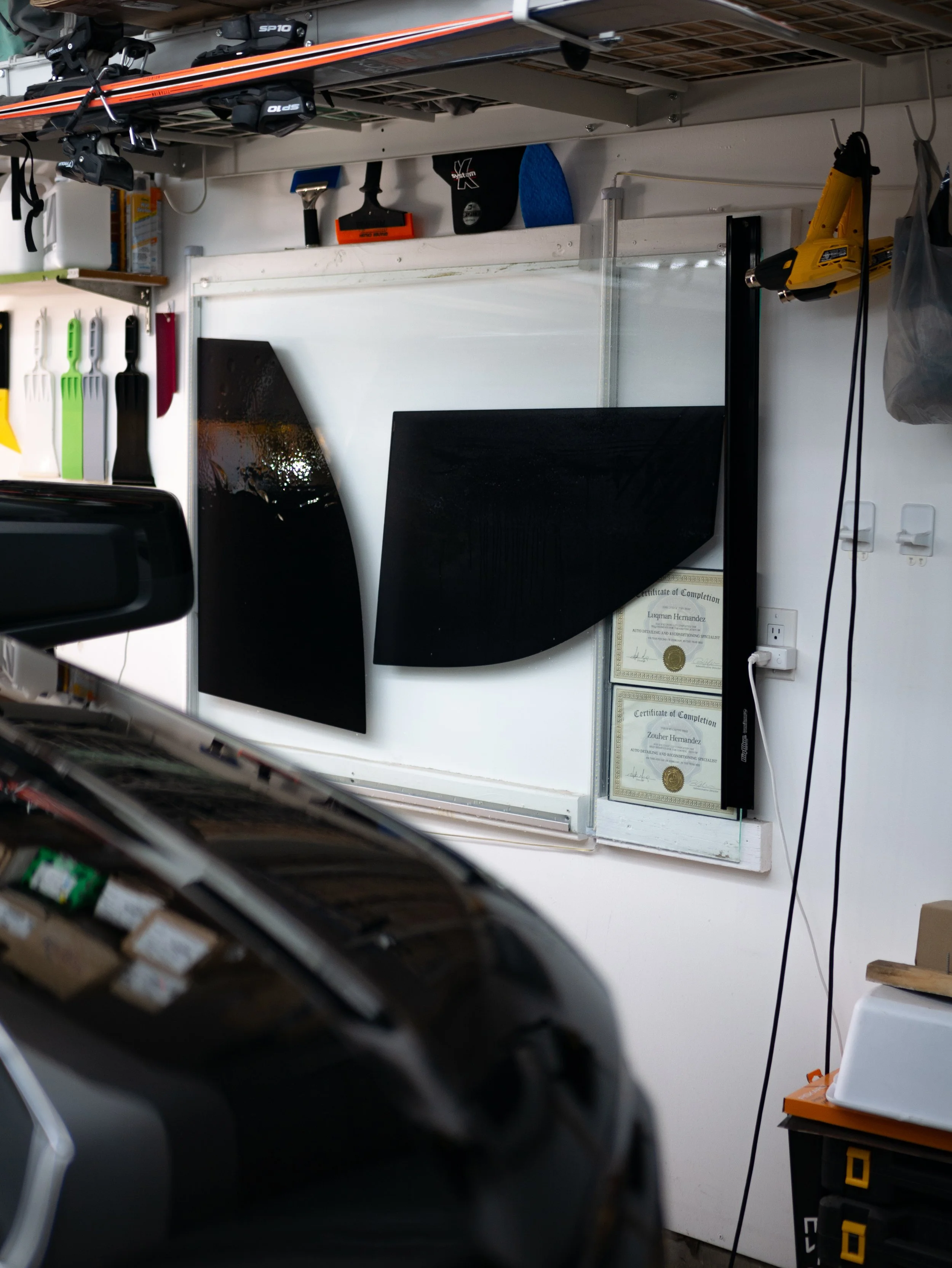 Garage or workshop with two black surfboard or skateboard fins mounted on the wall, various tools hanging including spatulas, a scraper, a black and orange squeegee, certificates on the wall, and a yellow power tool hanging on the right side.