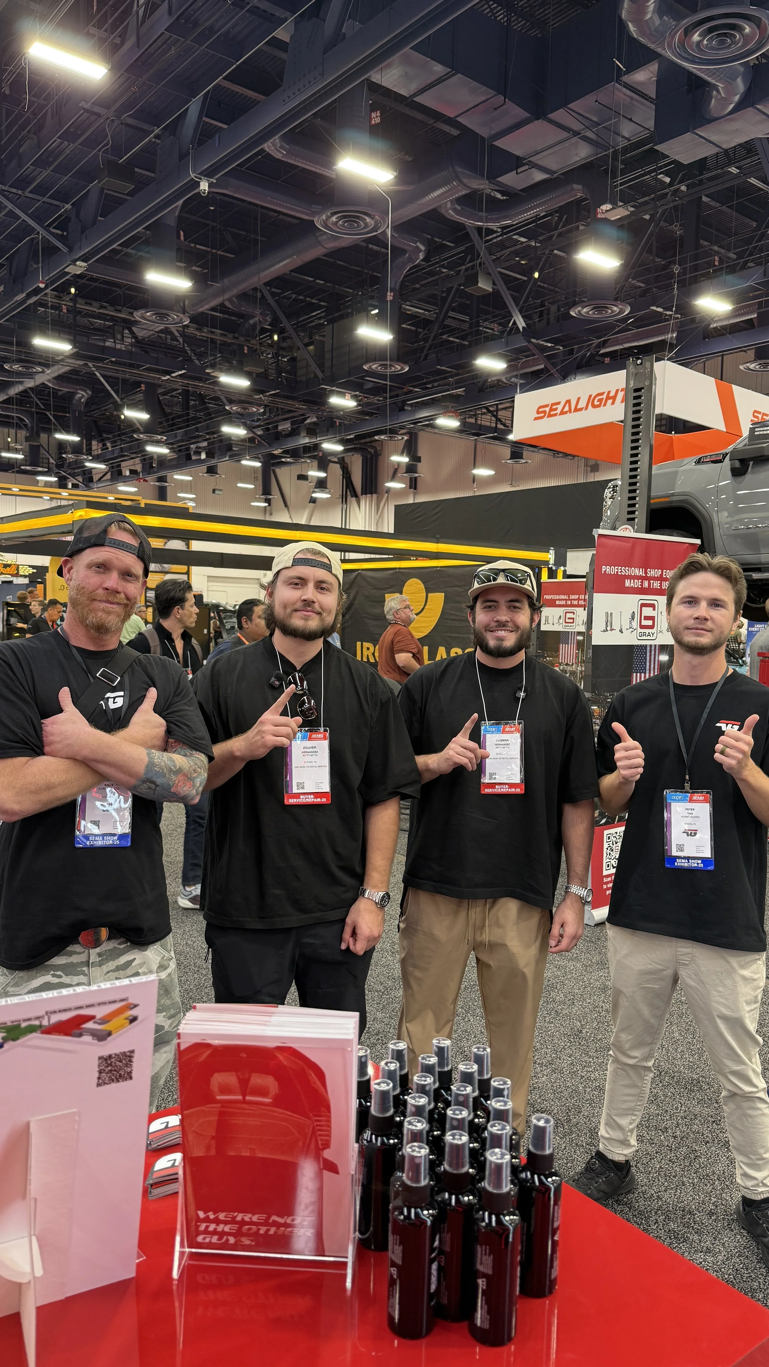 Group of four men at a trade show booth, posing with thumbs up and finger pointing, with product displays and signage in the background.