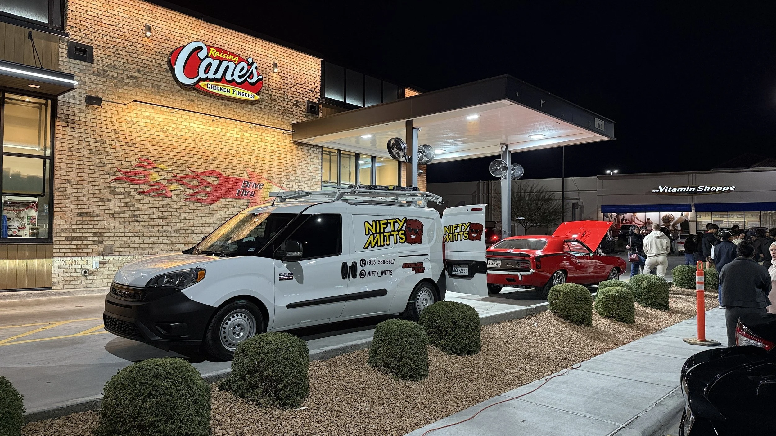 Night scene outside Raising Cane's restaurant with a white Nifty Mitts branded van parked beside a red car with its hood open, crowd of people near a shop called The Vitamin Shoppe, and small bushes along the sidewalk.