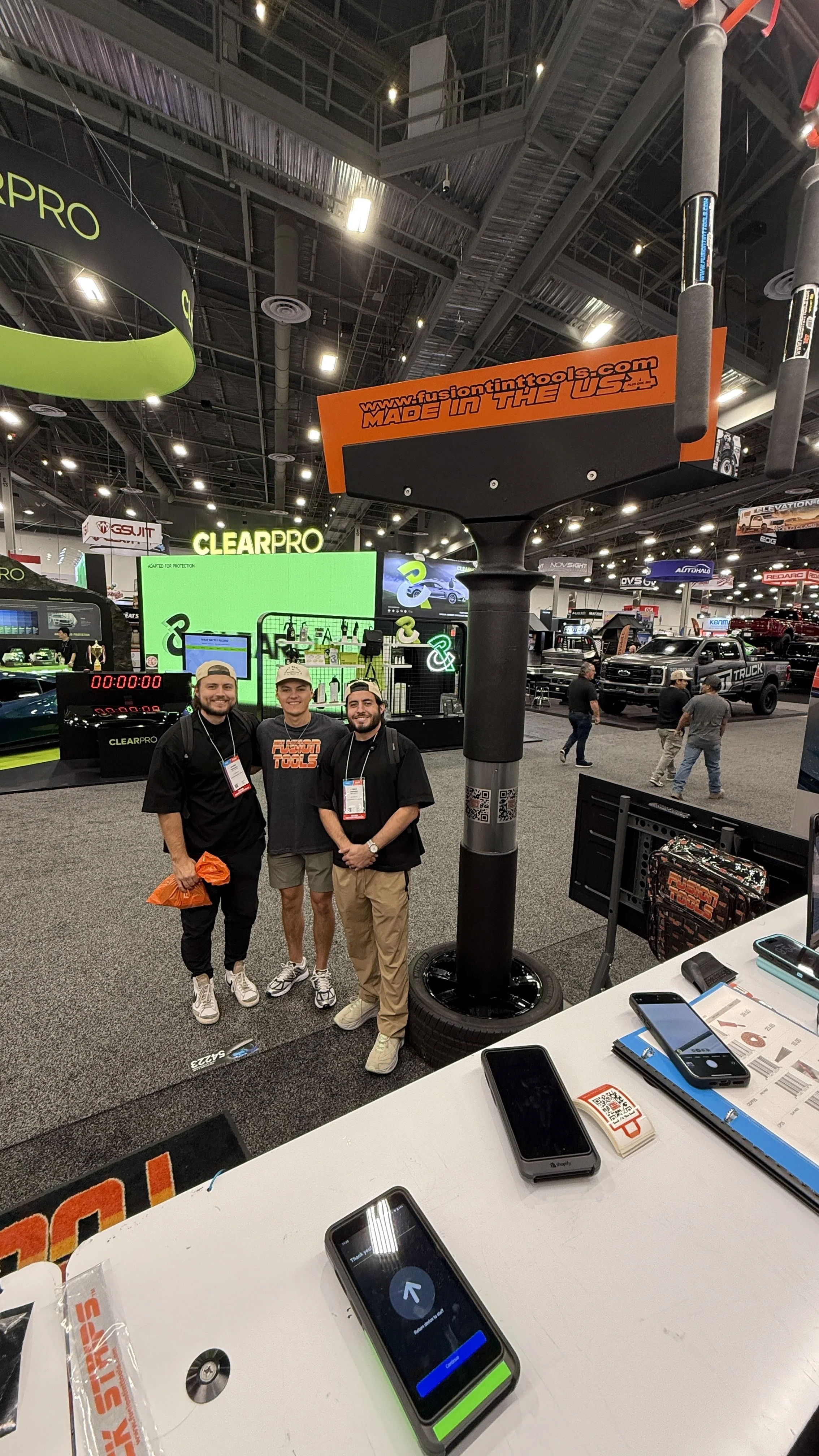 Three men standing together at a trade show booth with signs and displays for aftermarket automotive products, including a tall sign reading 'www.fusiontinntools.com MADE IN THE US,' and various electronic devices and smartphones on a table in the foreground.
