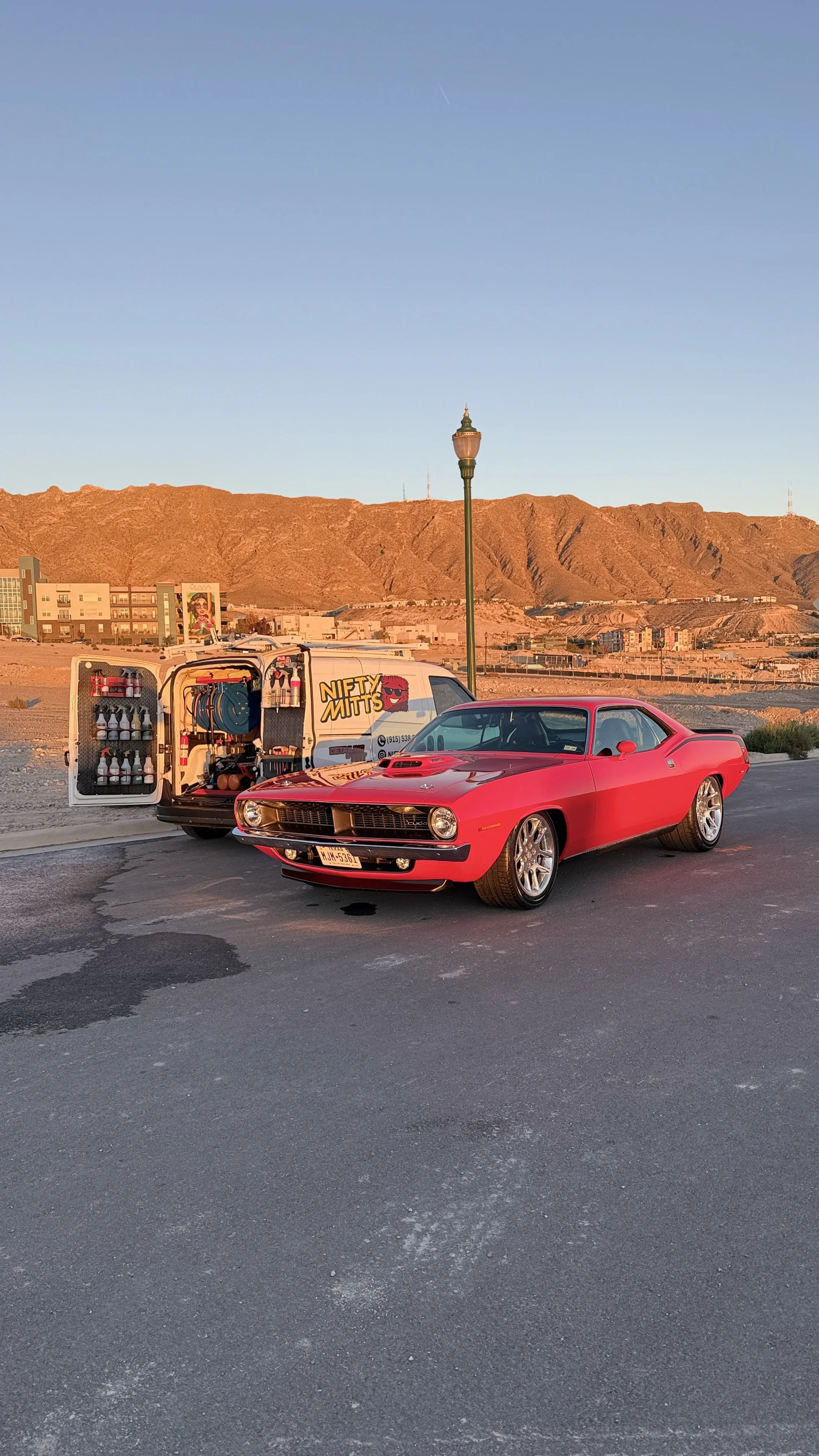 A red vintage muscle car parked on the street next to a white service van with equipment inside, in a desert area with mountains in the background, under a clear blue sky.