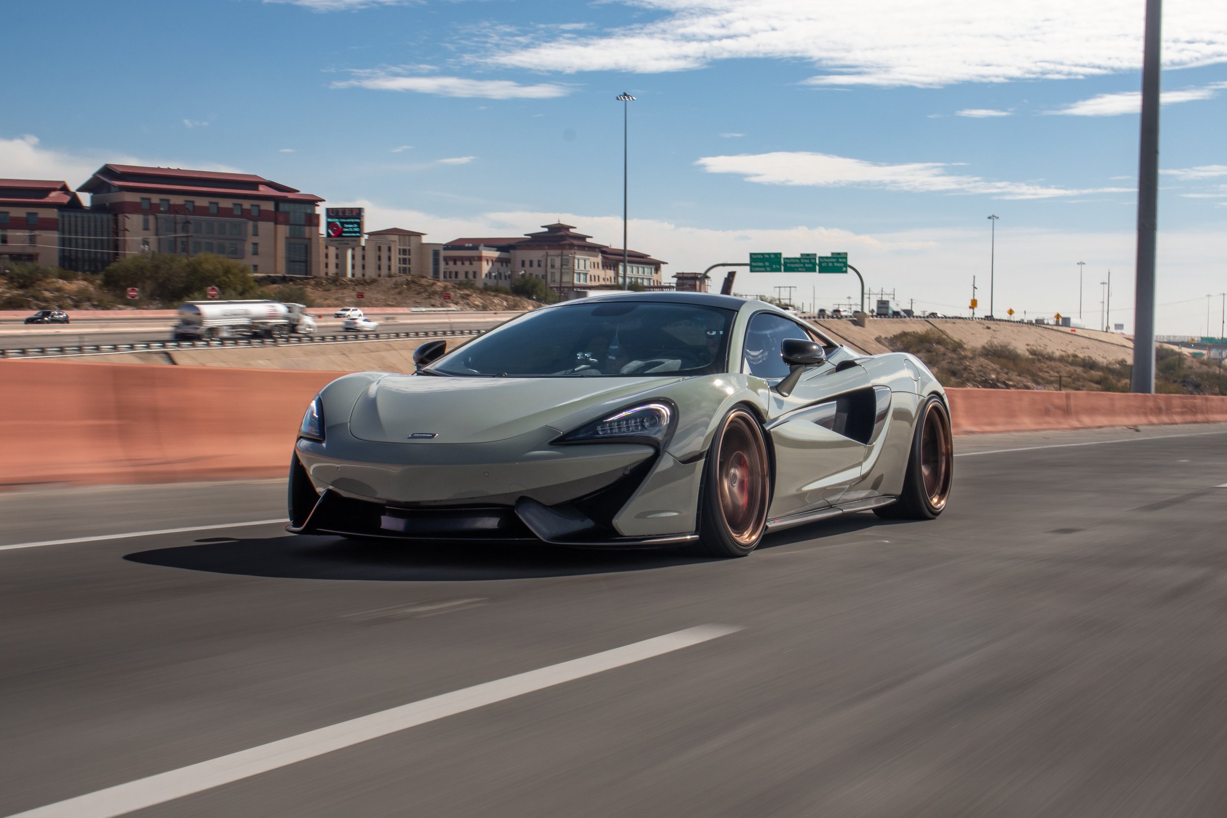 A sleek, silver sports car with dark bronze rims driving on a highway under a partly cloudy sky, with buildings and other vehicles in the background.
