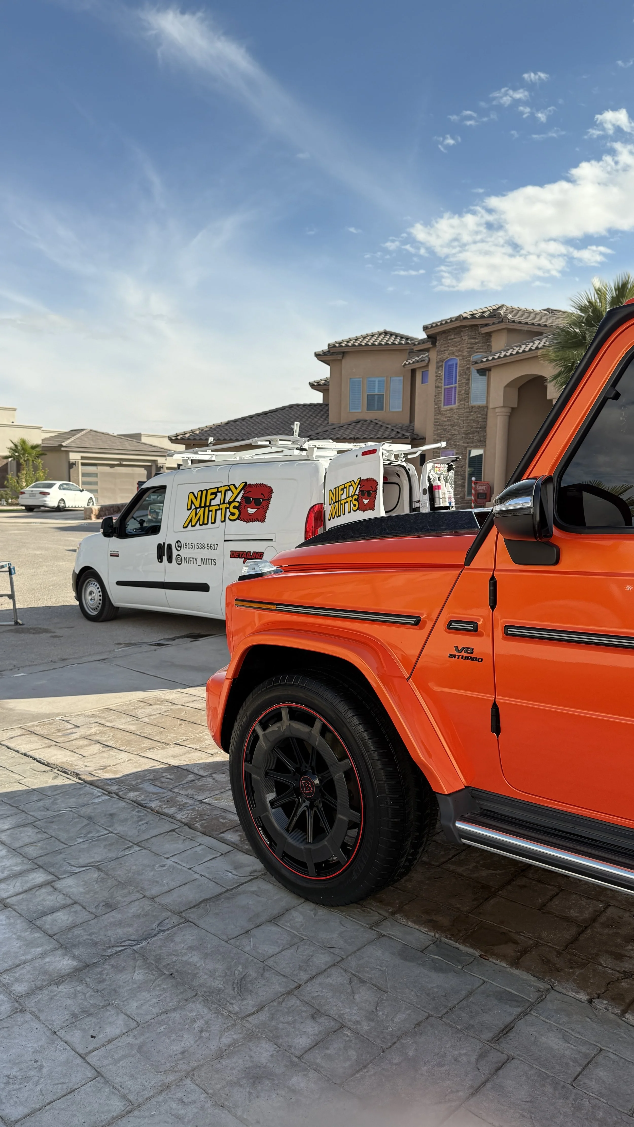 View of a white van with 'Nifty Mitts' branding and a cartoon face logo, parked next to an orange luxury SUV in a residential neighborhood under a blue sky with clouds.