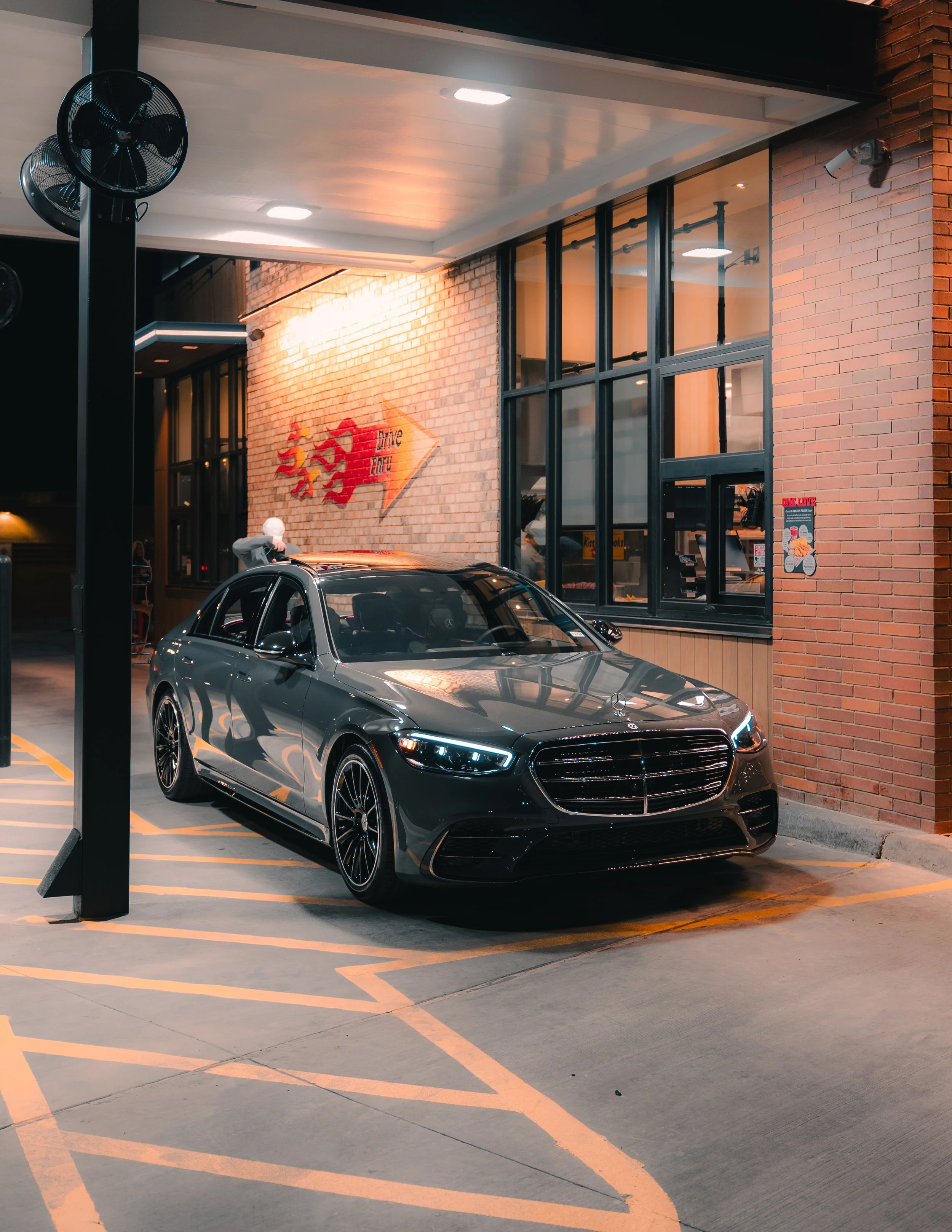 A black Mercedes-Benz sedan parked under a covered drive-thru at night outside a restaurant with a brick exterior and large windows.