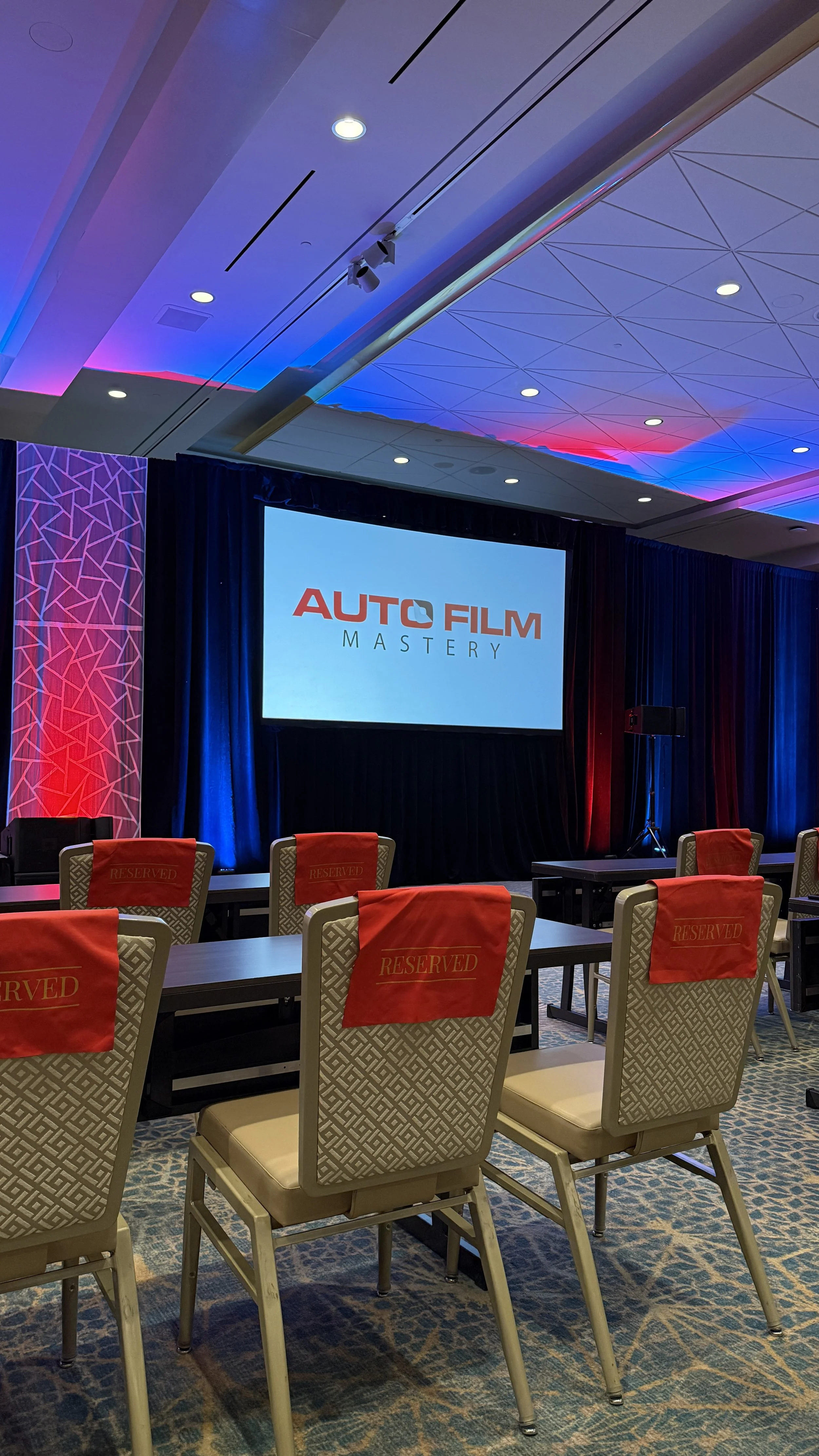 Conference room with chairs reserved with red cloths, a large screen displaying 'Auto Film Mastery,' and stage lighting in red and blue.