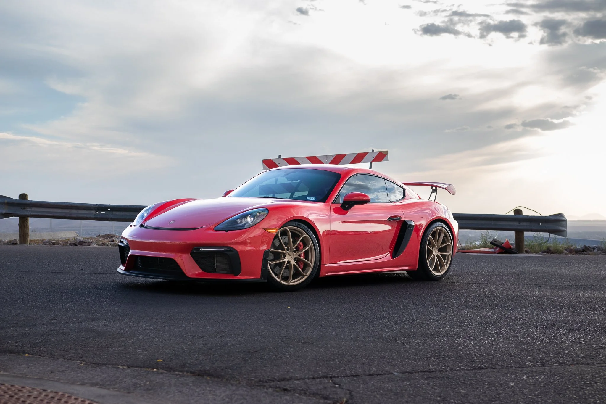 A red sports car with a rear spoiler and black accents parked on a paved road at sunset, with a guardrail and cloudy sky in the background.