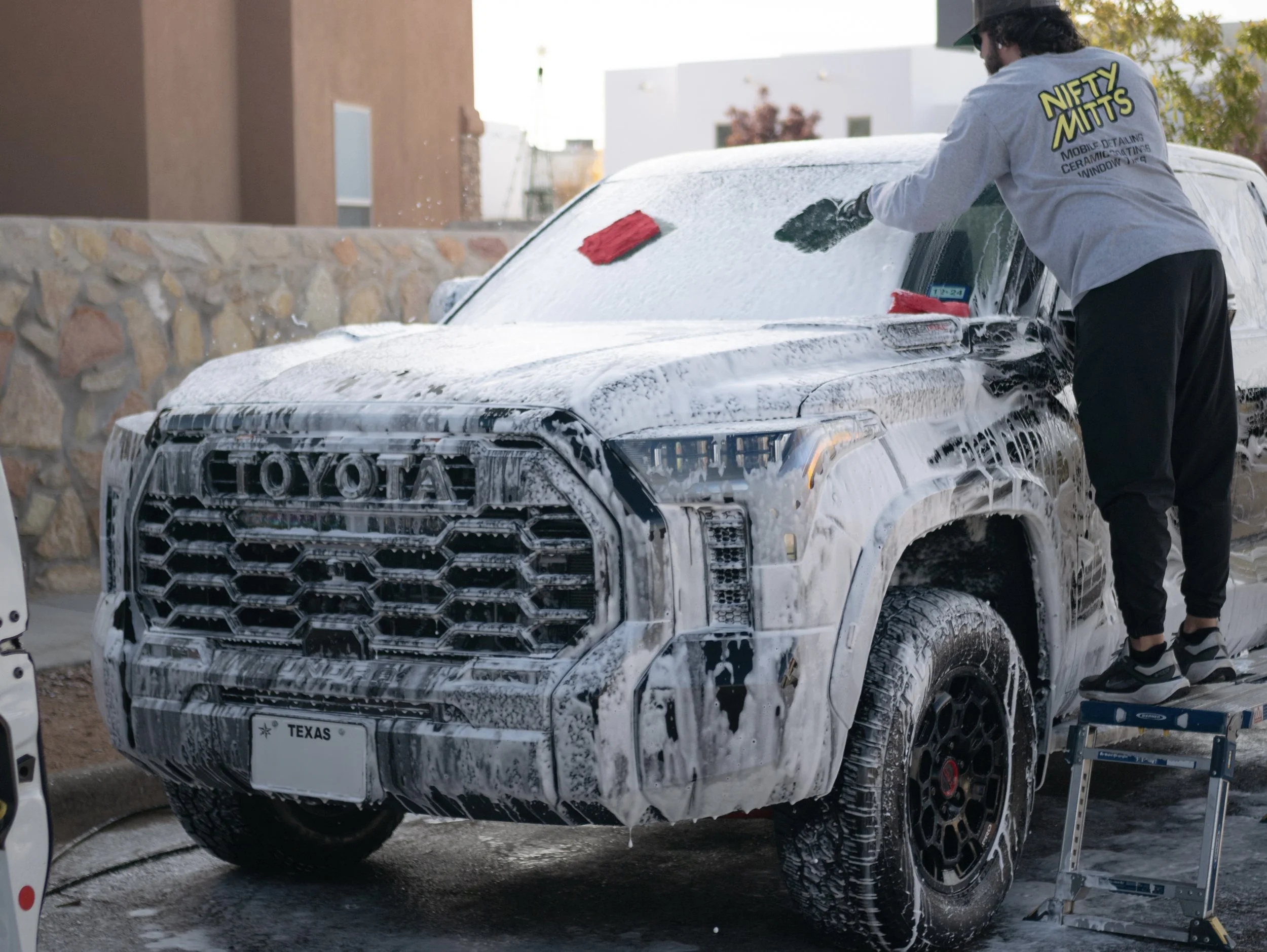 Man washing a black Toyota SUV with soap and a brush while standing on a small step ladder. The SUV is covered in soap suds, and the background shows urban buildings and a stone wall.