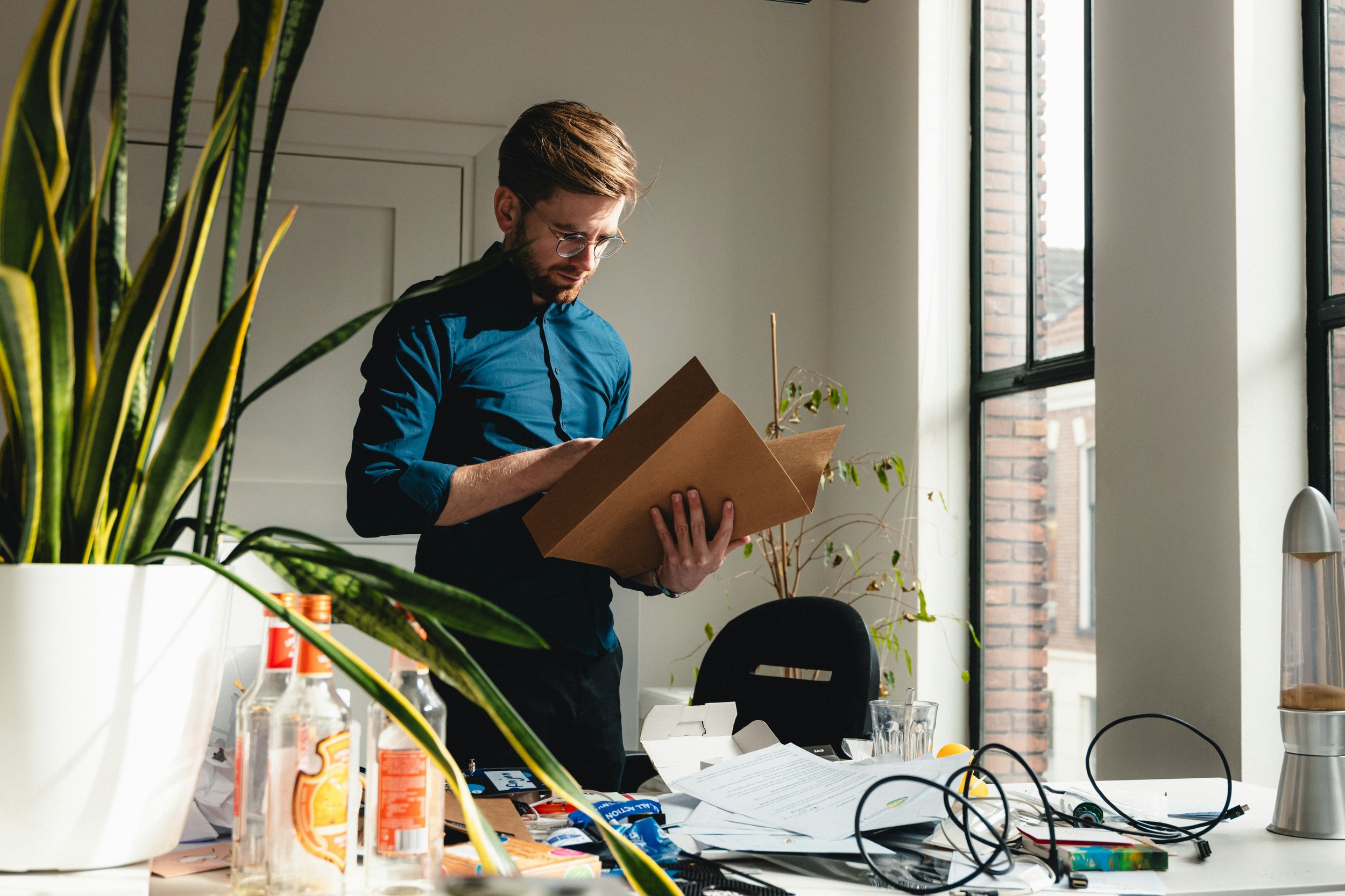 A man with brown hair and glasses wearing a blue shirt, standing indoors by large windows, reading a brown cardboard box surrounded by clutter and plants.