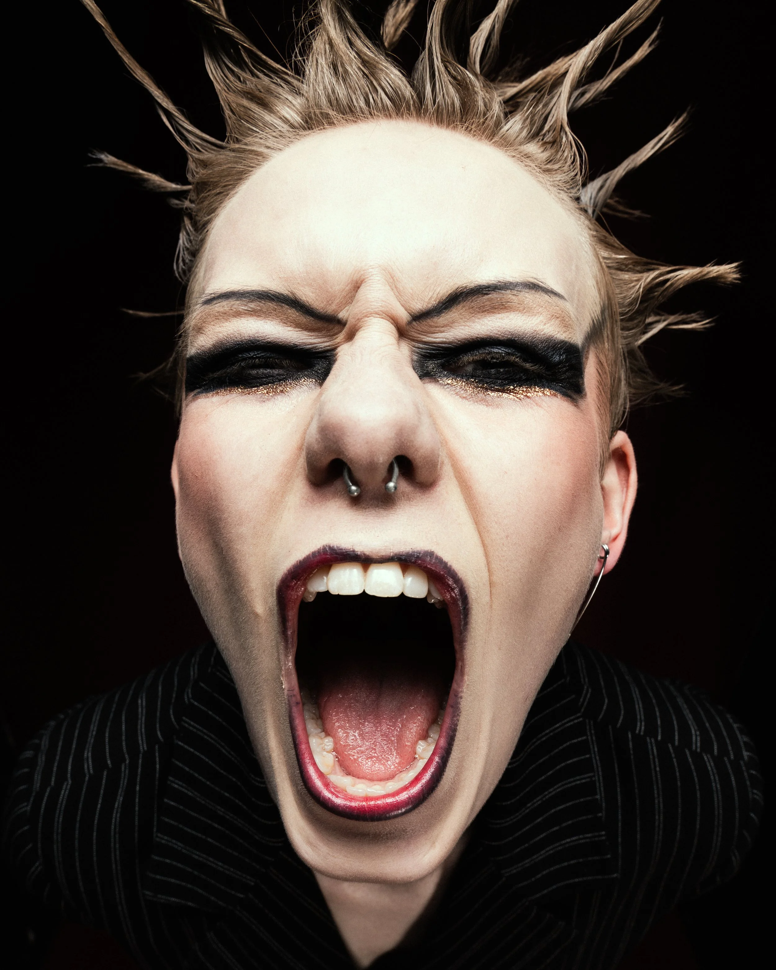 A person with spiky blonde hair, black eye makeup, and a septum piercing, screaming with their mouth wide open, wearing a striped shirt, against a black background.