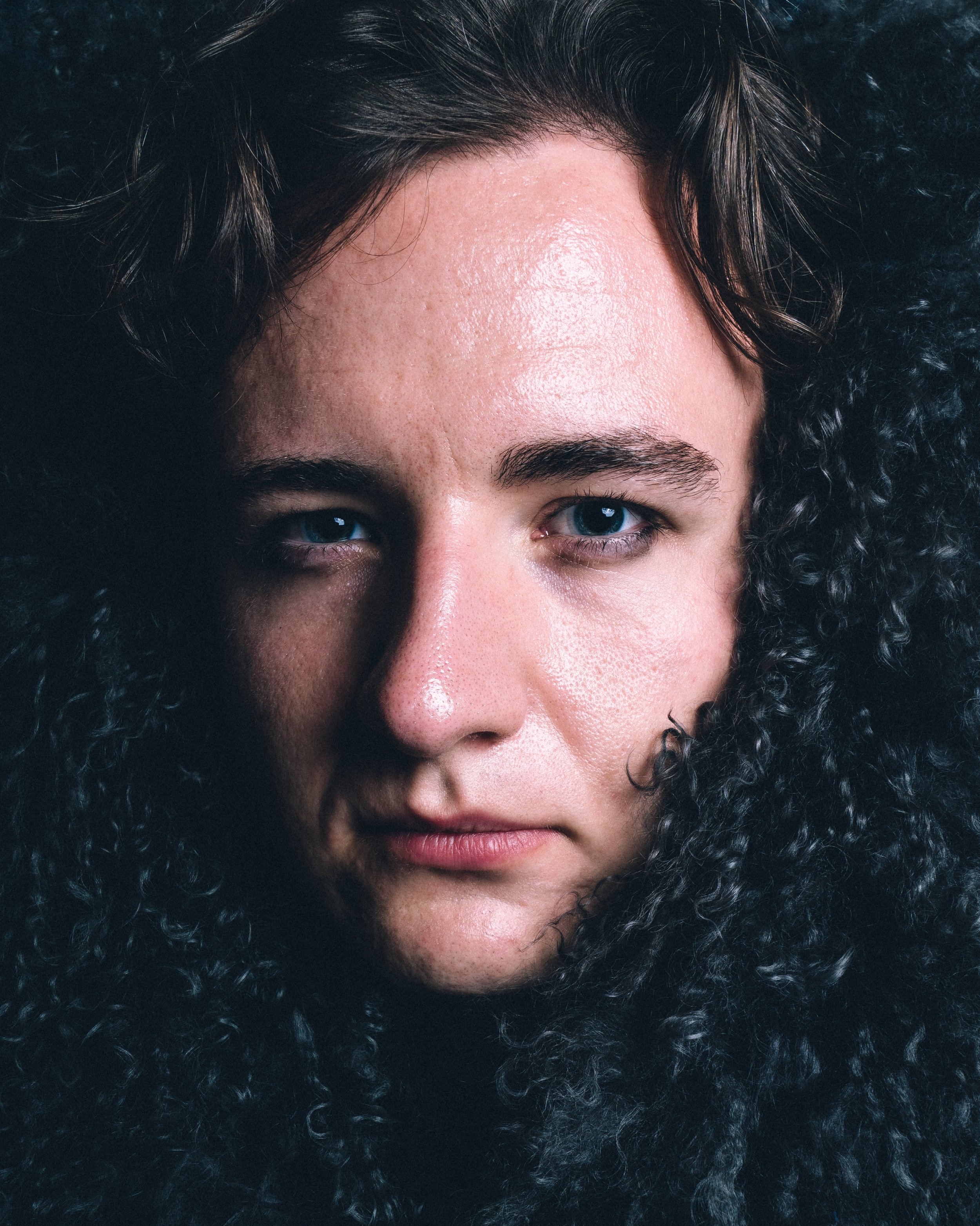 Close-up portrait of a person with long, curly black hair and piercing blue eyes.