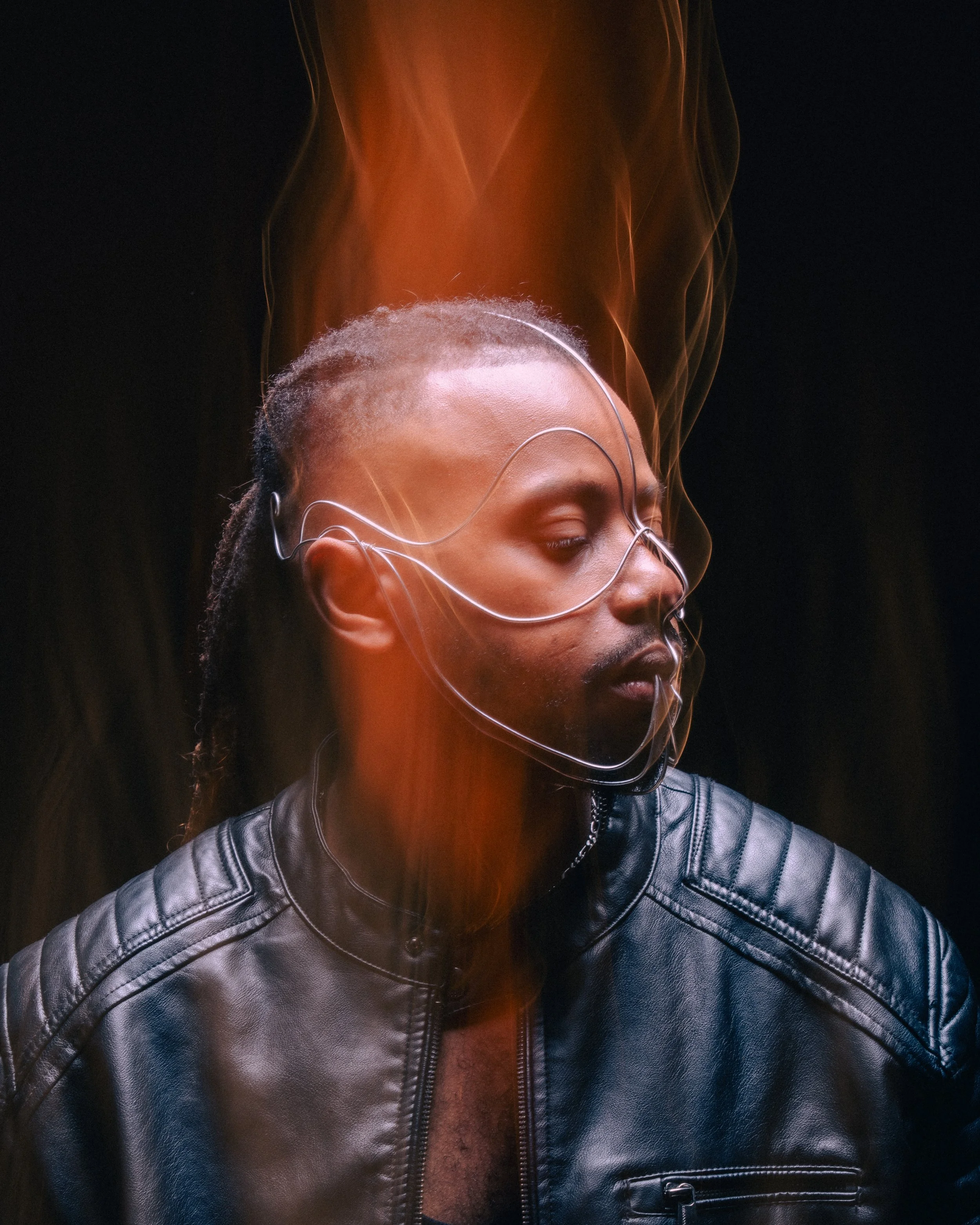 A person with dreadlocks wearing a leather jacket and large, futuristic glasses has their eyes closed as vibrant orange light and smoke swirl around their head.