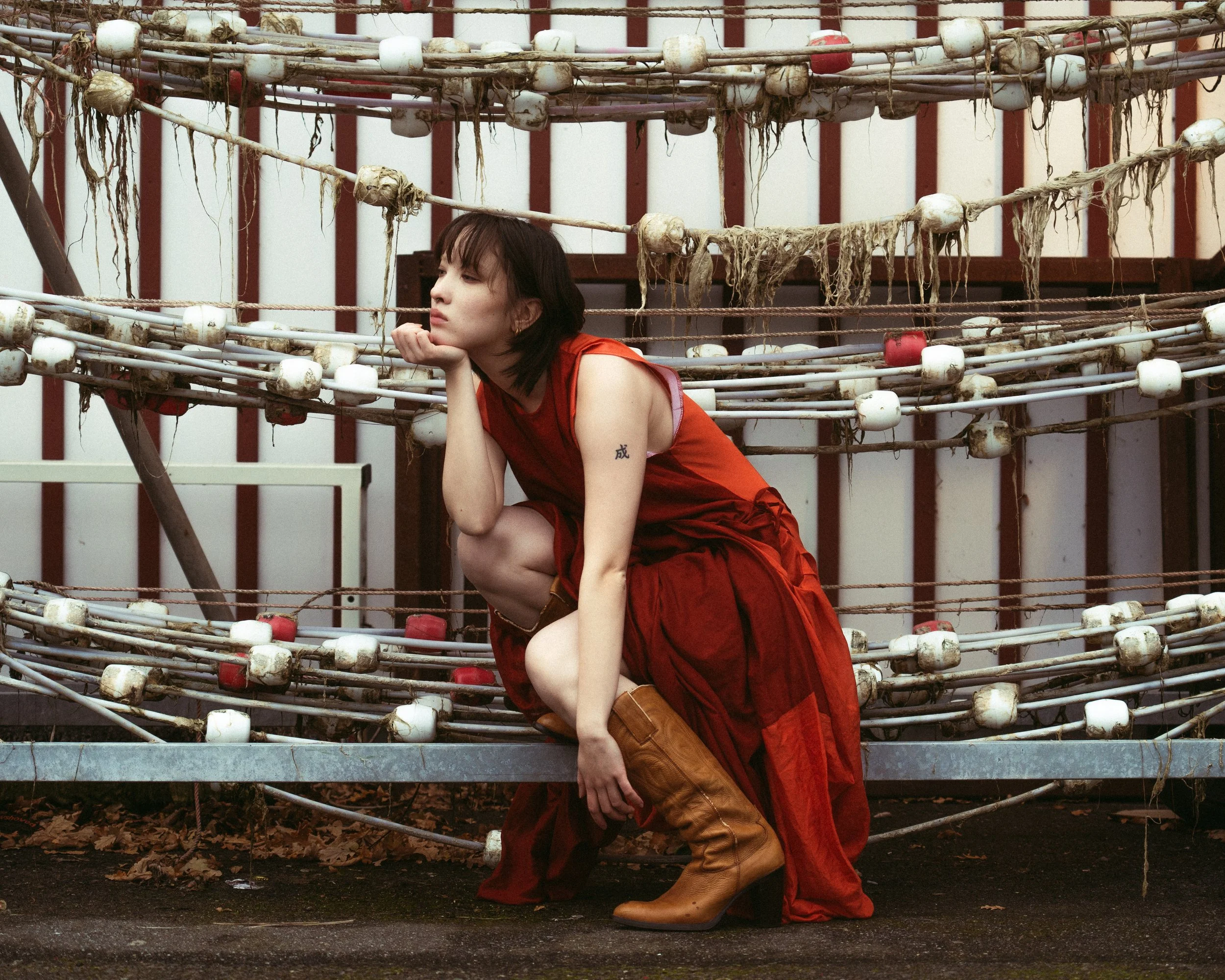 A woman in a red dress and tan boots crouches in front of an old, rusted carnival ride with worn, tangled ropes and plastic balls, looking contemplative.