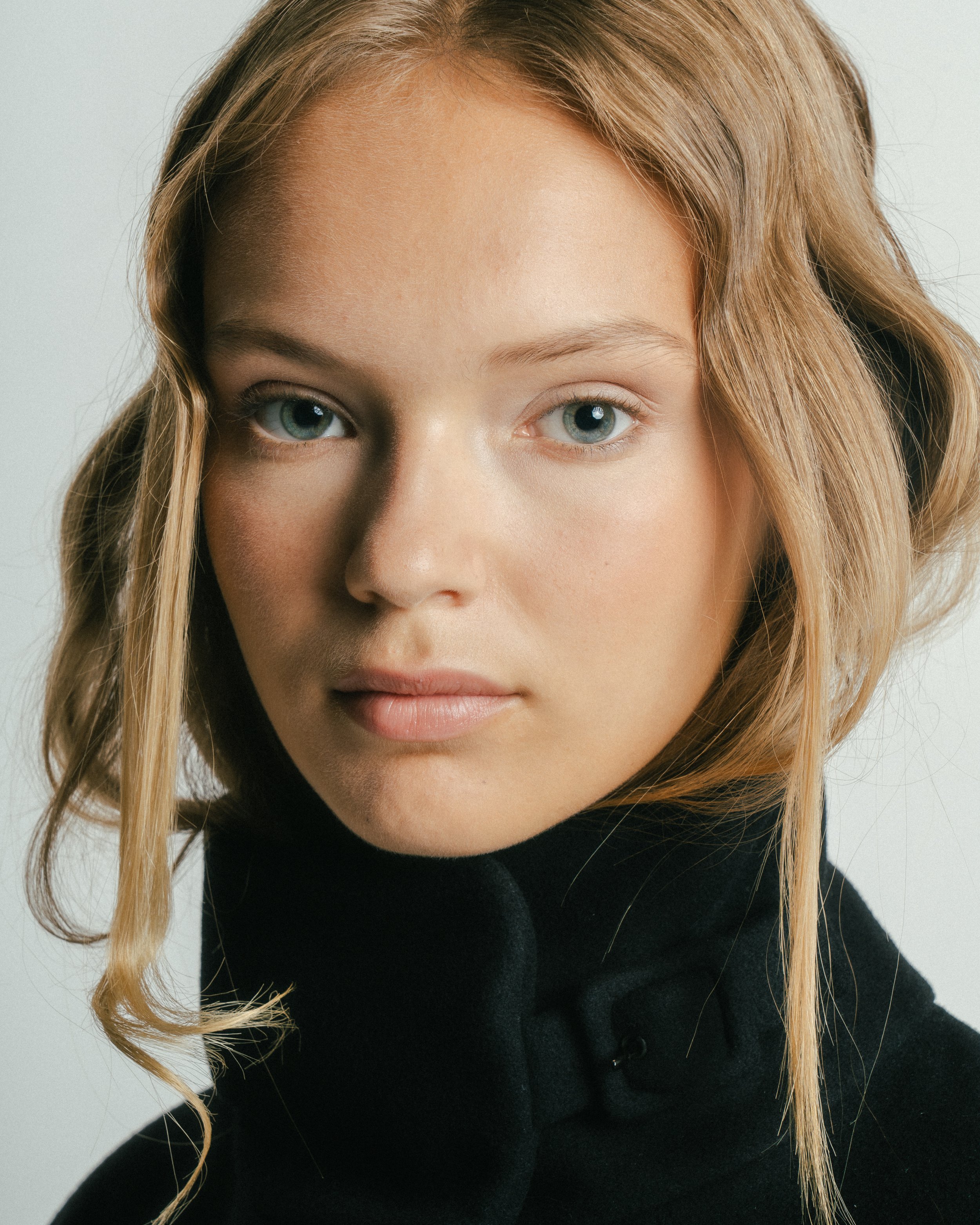 Close-up portrait of a young woman with blonde hair, blue eyes, and fair skin, wearing a black turtleneck sweater.
